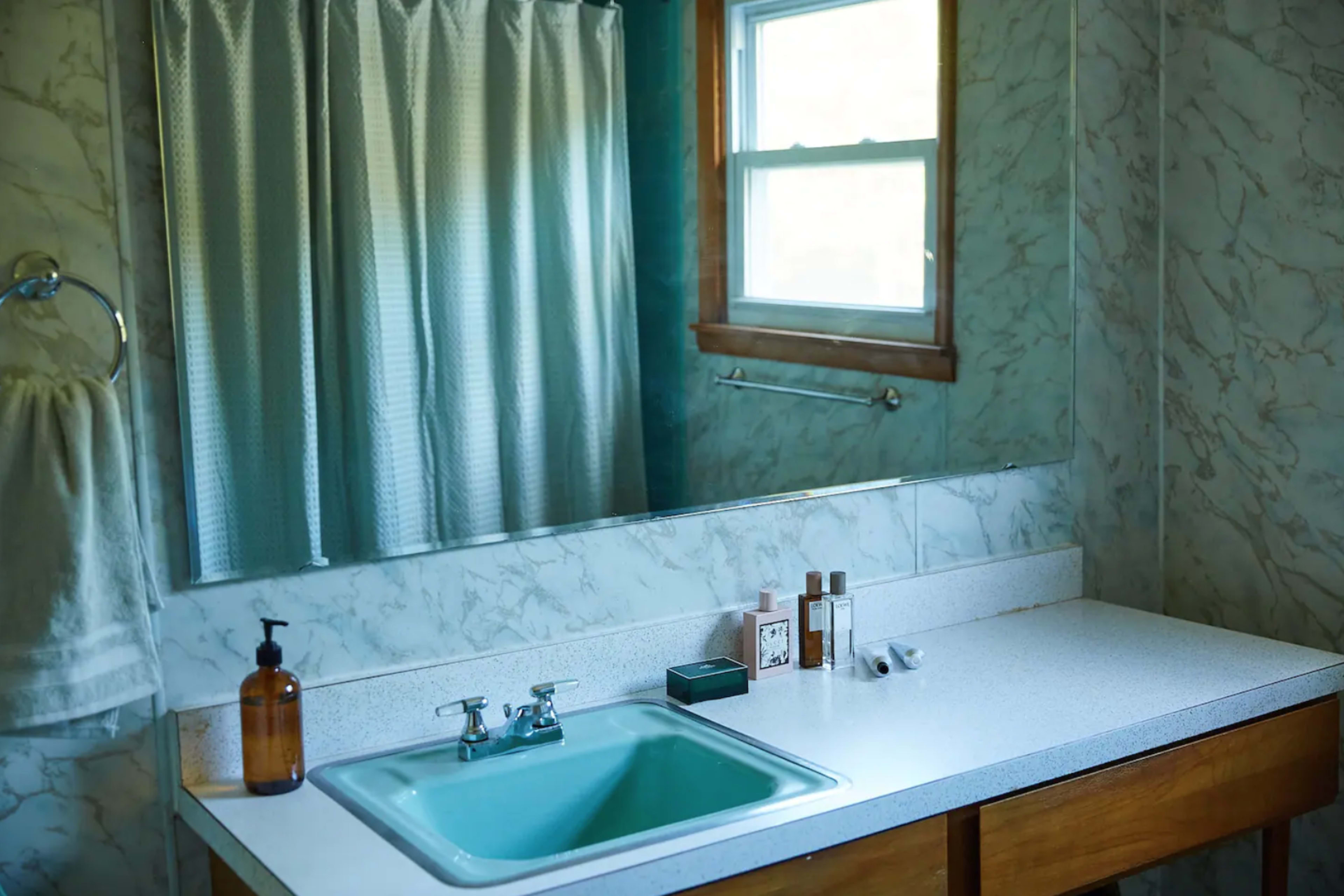 A bathroom with a green sink, a mirror, a window, and a shower curtain, along with various toiletries placed on the countertop.