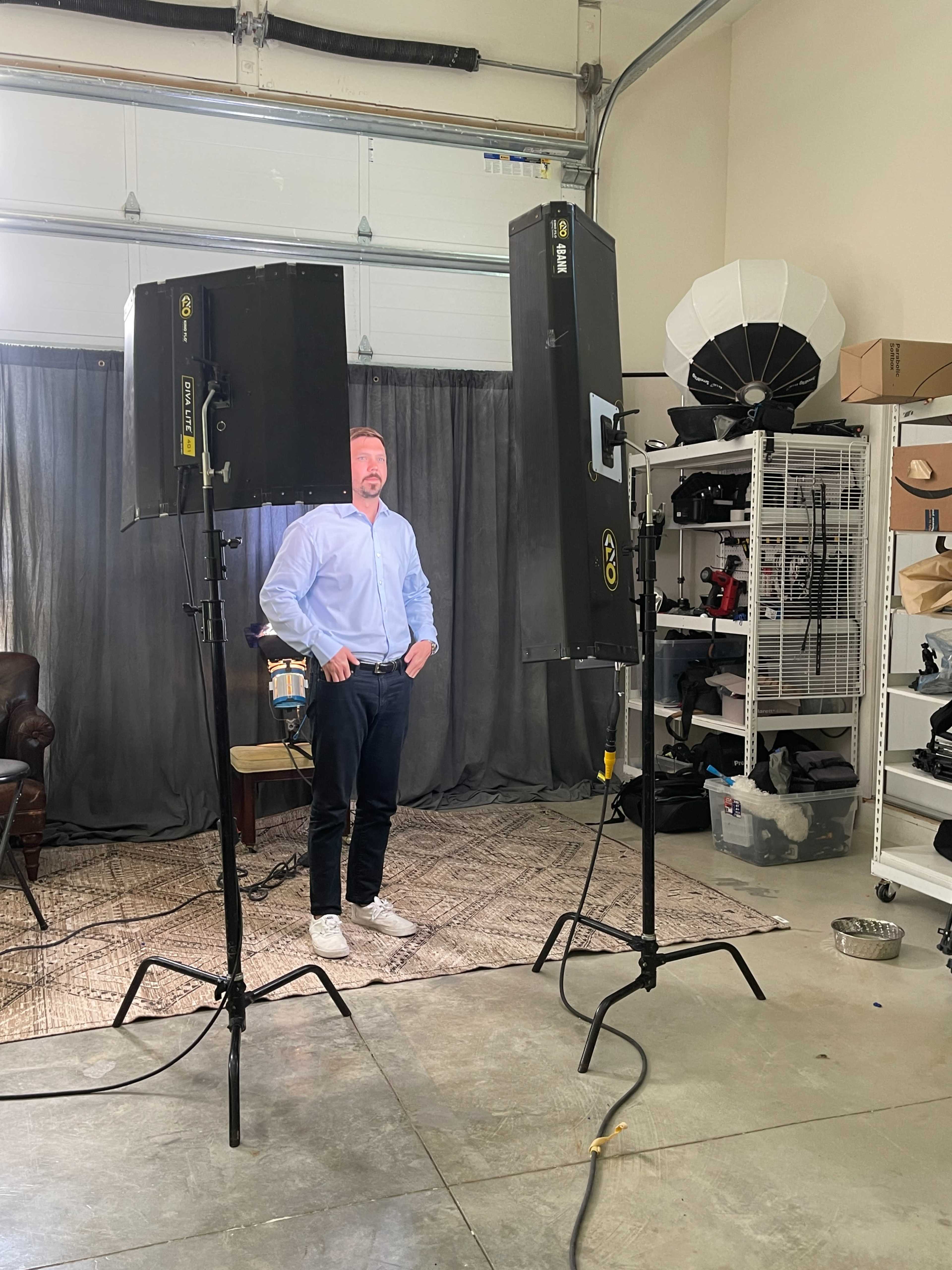 A man in a blue shirt stands between two lighting setups in a studio space with a grey backdrop.