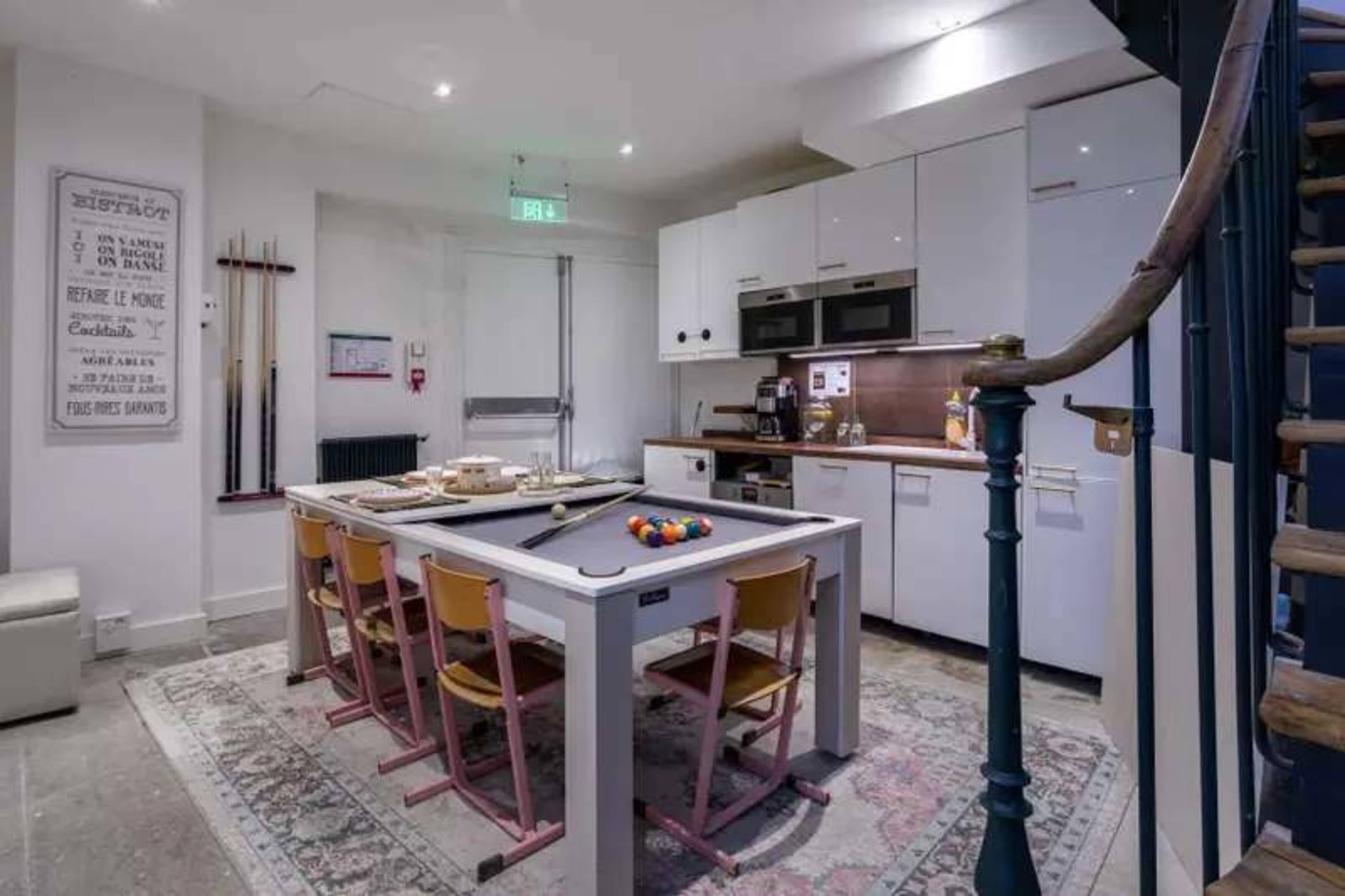 The image shows a modern kitchen with a pool table in the center, surrounded by four yellow chairs, and features a spiral staircase in the background.