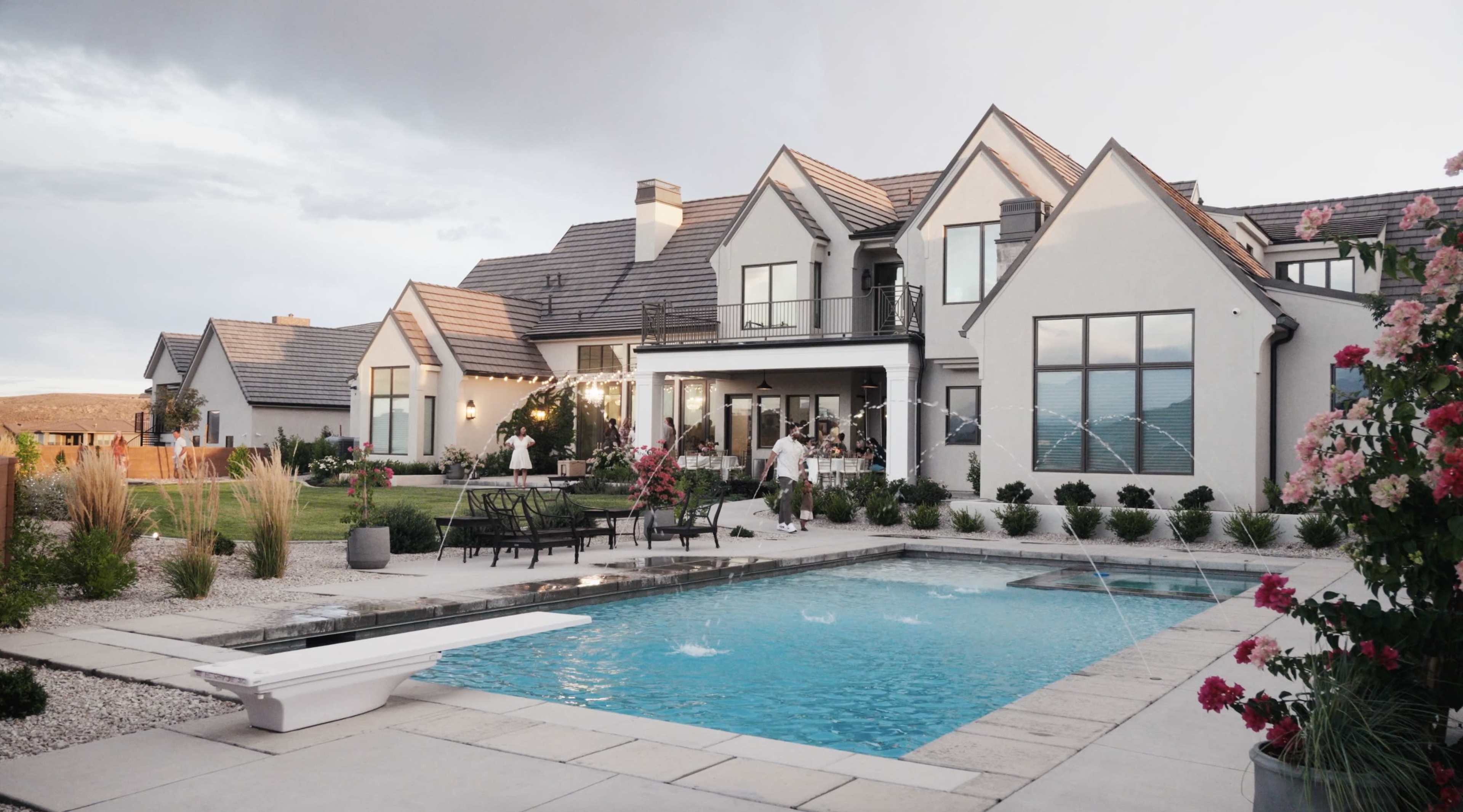 A modern house with multiple gabled roofs surrounds a rectangular swimming pool, flanked by a landscaped yard and patio furniture.