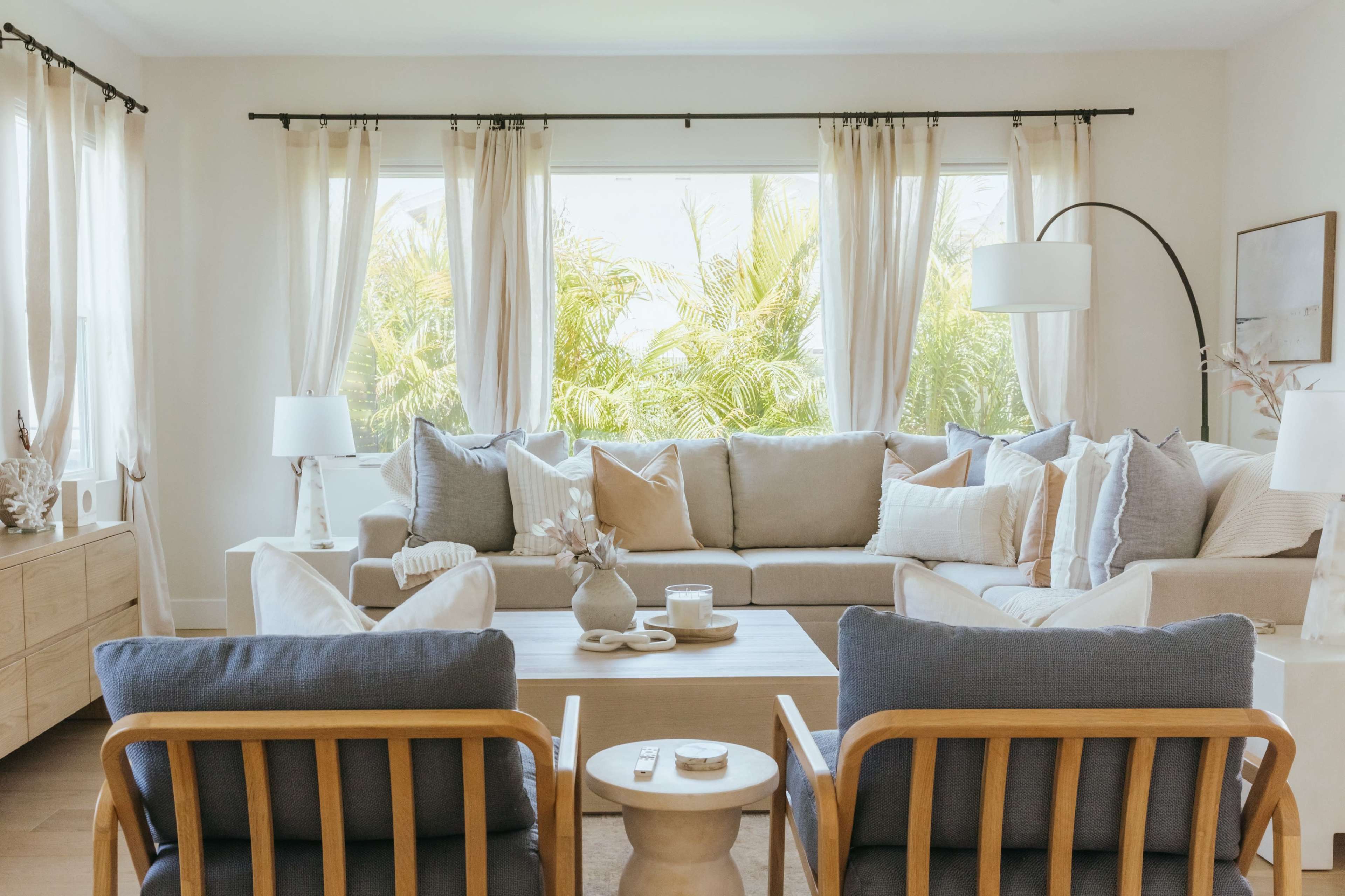 A bright living room features a light-colored sofa surrounded by armchairs, coffee table, and large windows with sheer curtains offering a view of greenery.