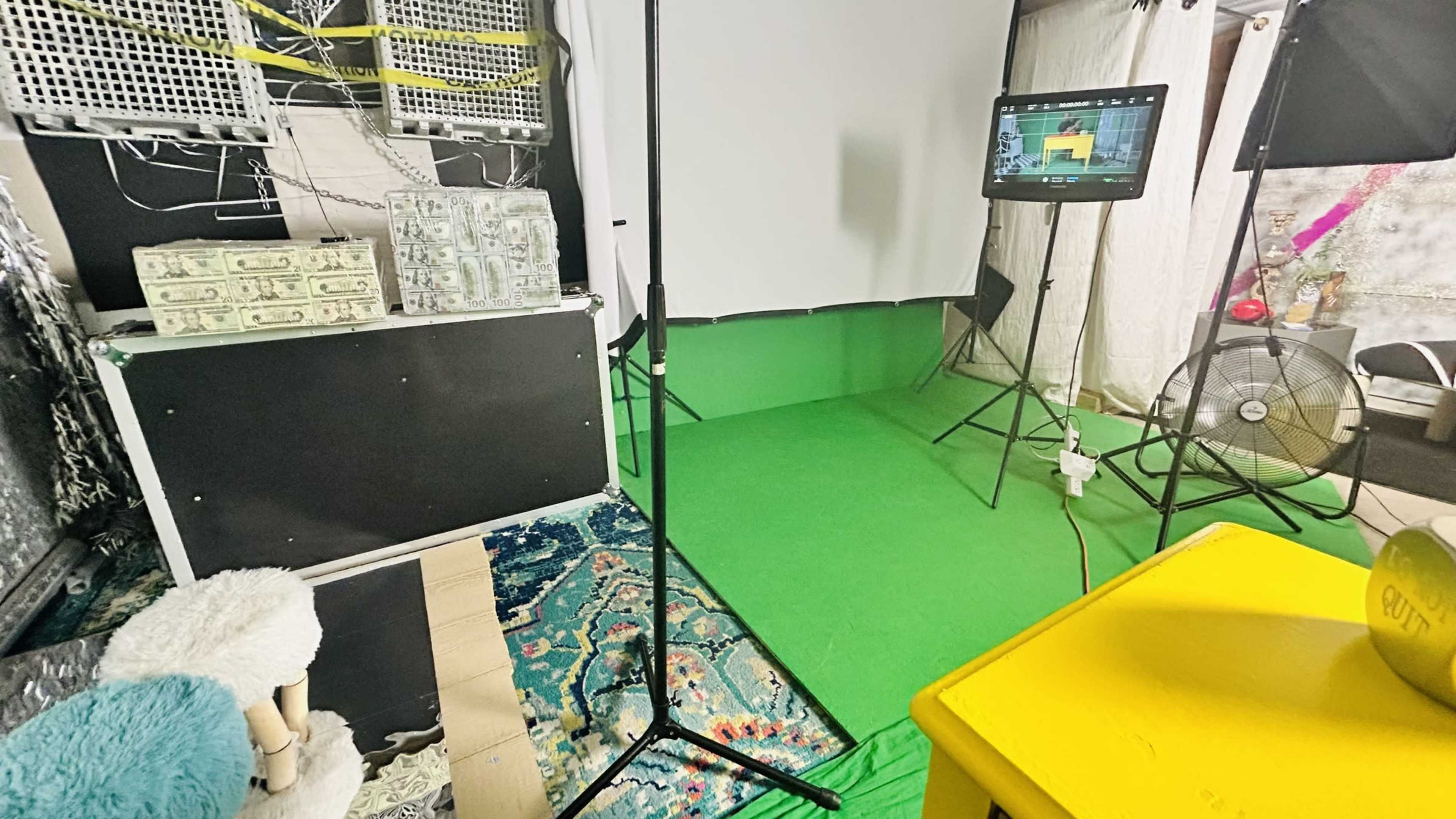 A studio setup featuring a green screen, a display monitor, lighting equipment, and a patterned rug on the floor.