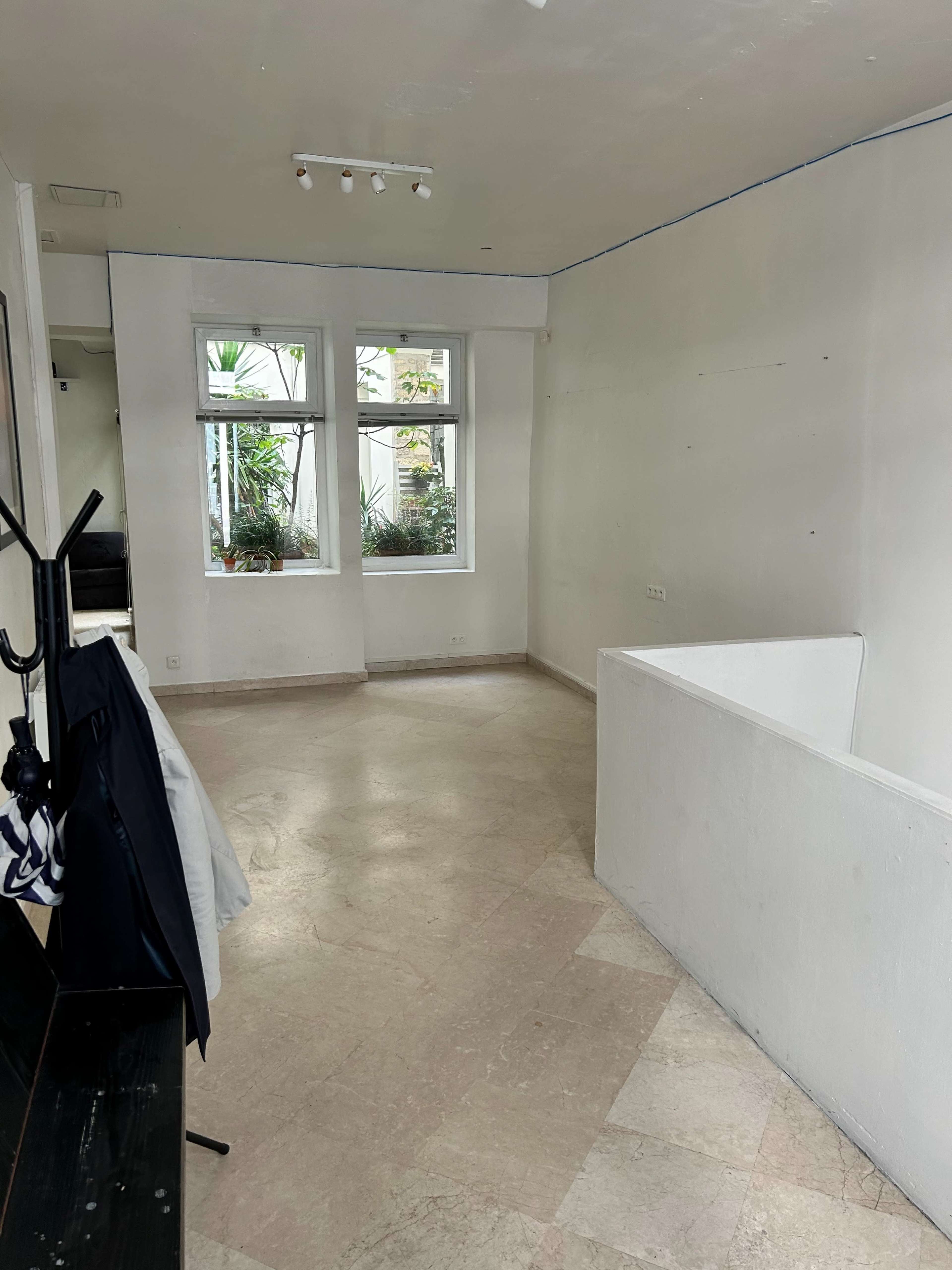 The image shows a spacious, empty room with light-colored walls and a tiled floor, featuring two windows with plants visible outside.