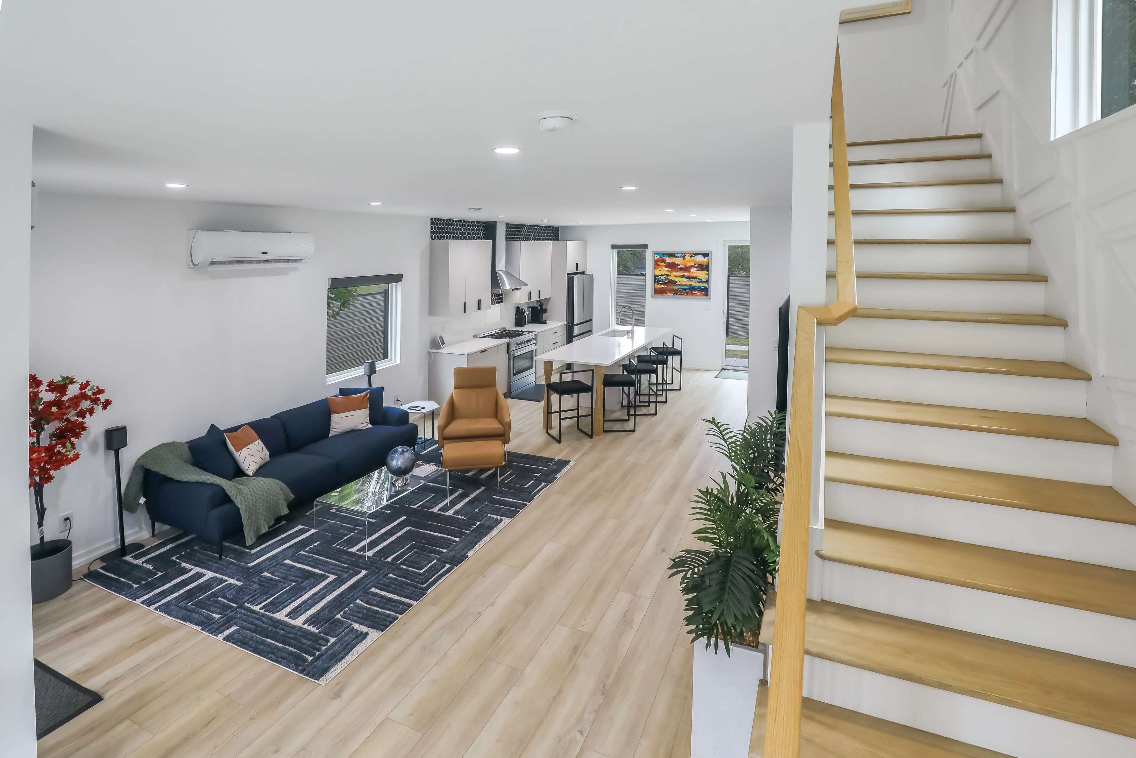 A modern open-concept living space with a staircase, featuring a blue sofa, a glass coffee table, a dining area, and large windows.