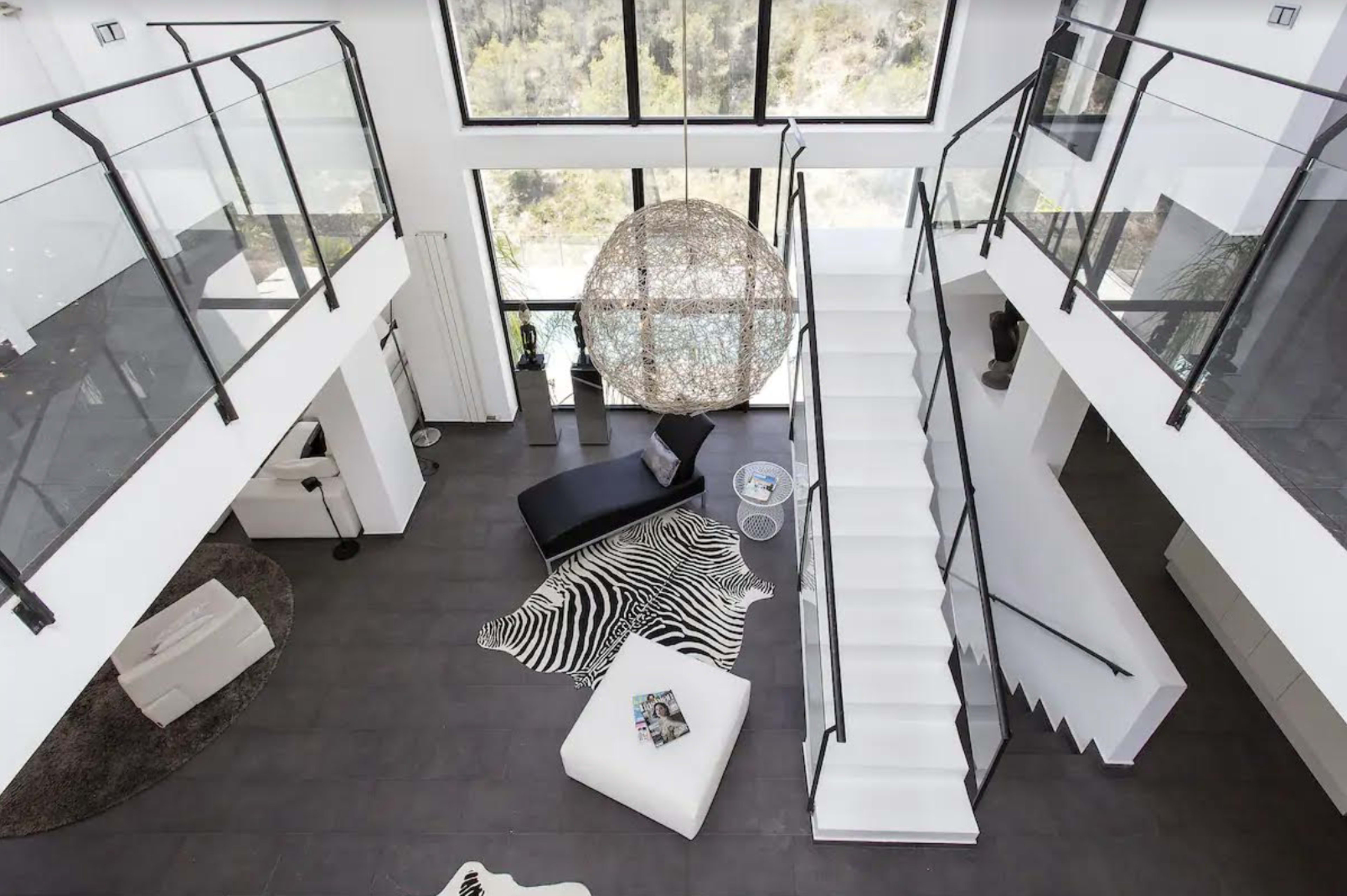A modern two-story living space with large windows, a curved staircase, and a round light fixture, featuring minimalist furniture and a patterned rug.