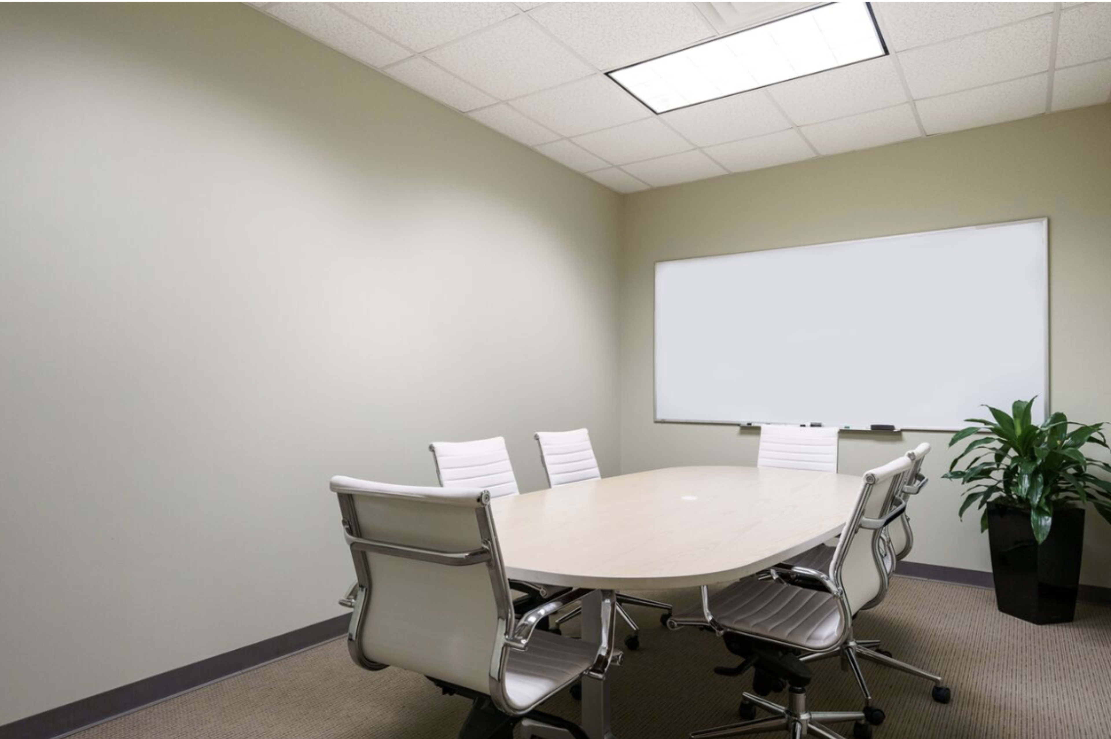 A conference room with a large oval table surrounded by six white chairs, a whiteboard on the wall, and a potted plant in the corner.