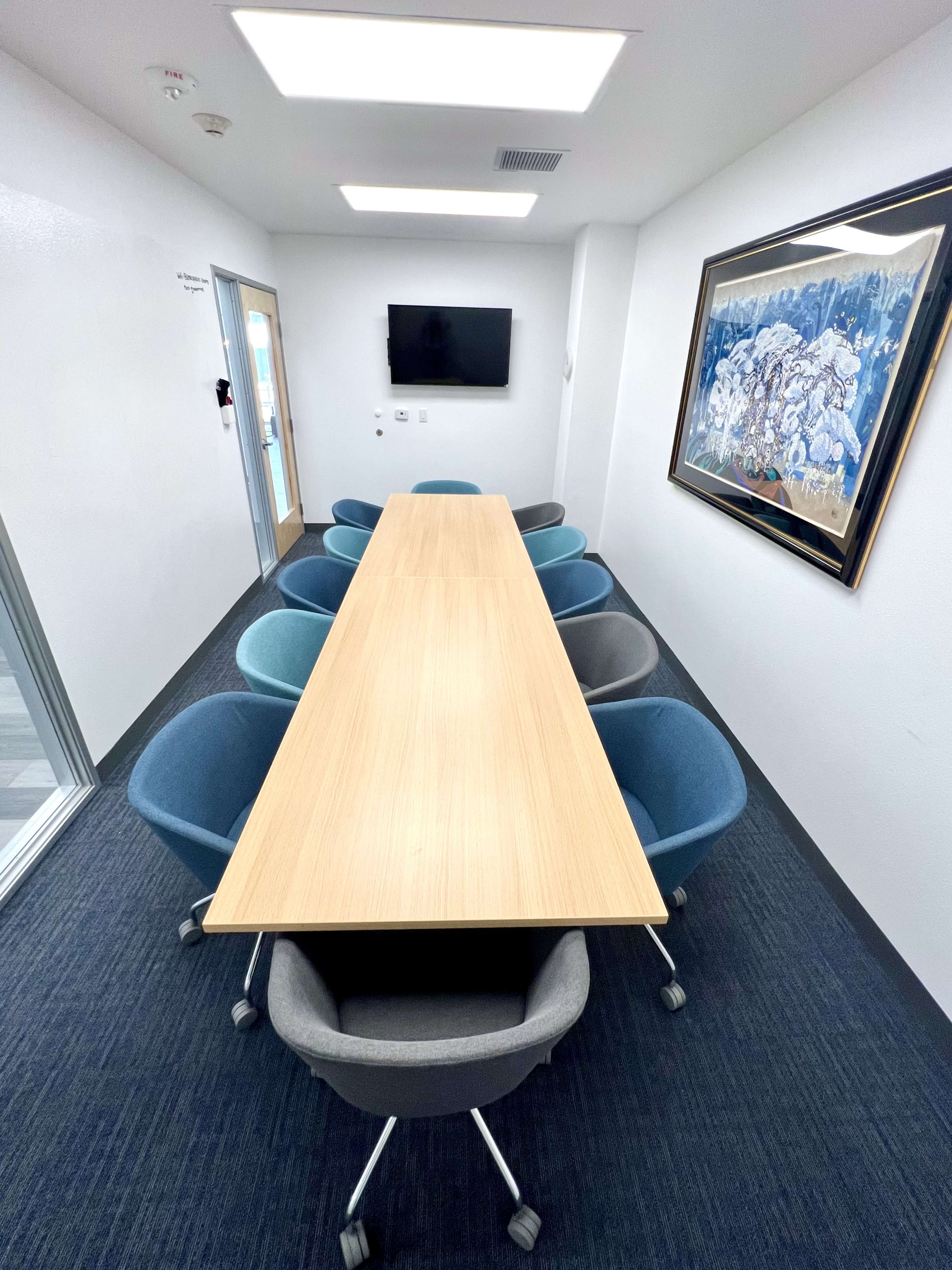A long wooden table surrounded by eight chairs is positioned in a bright meeting room with a television on the wall and a decorative artwork.