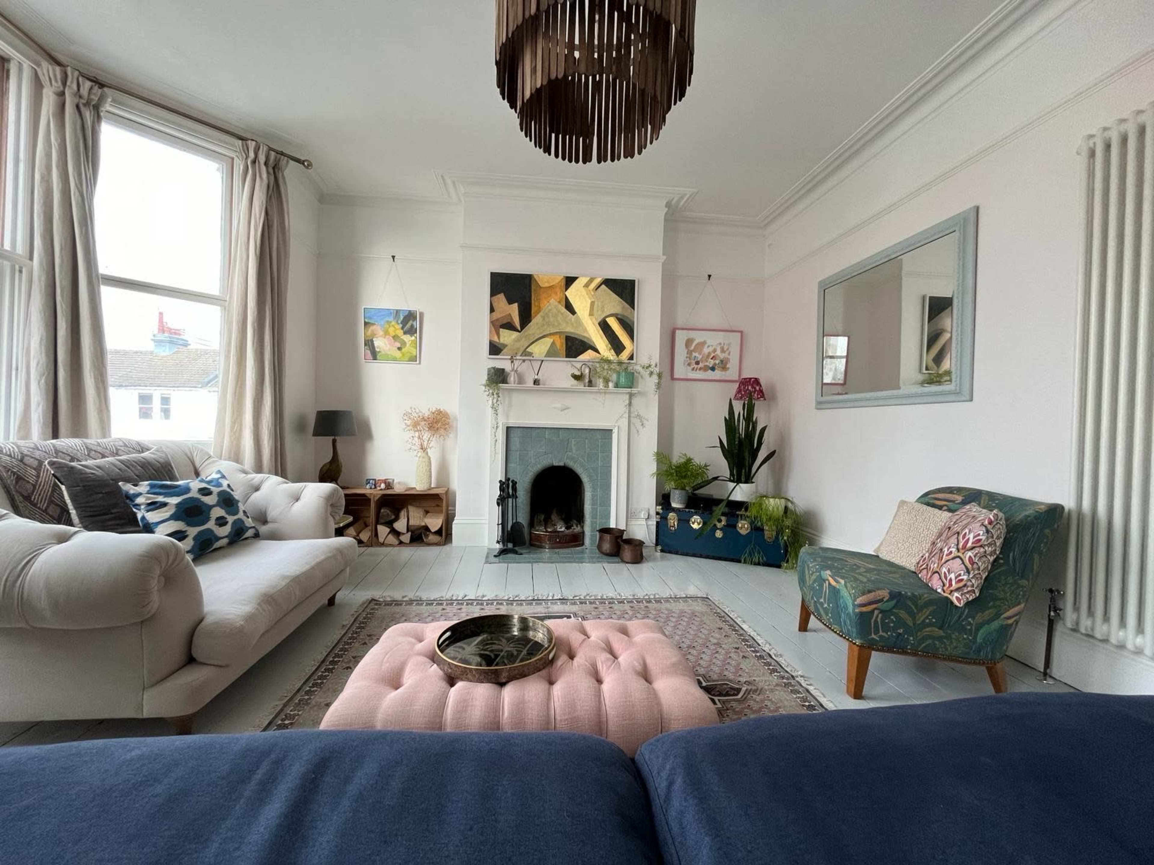 The living room features a cream sofa, a patterned armchair, a pink tufted coffee table, and a decorative fireplace surrounded by plants and artwork.