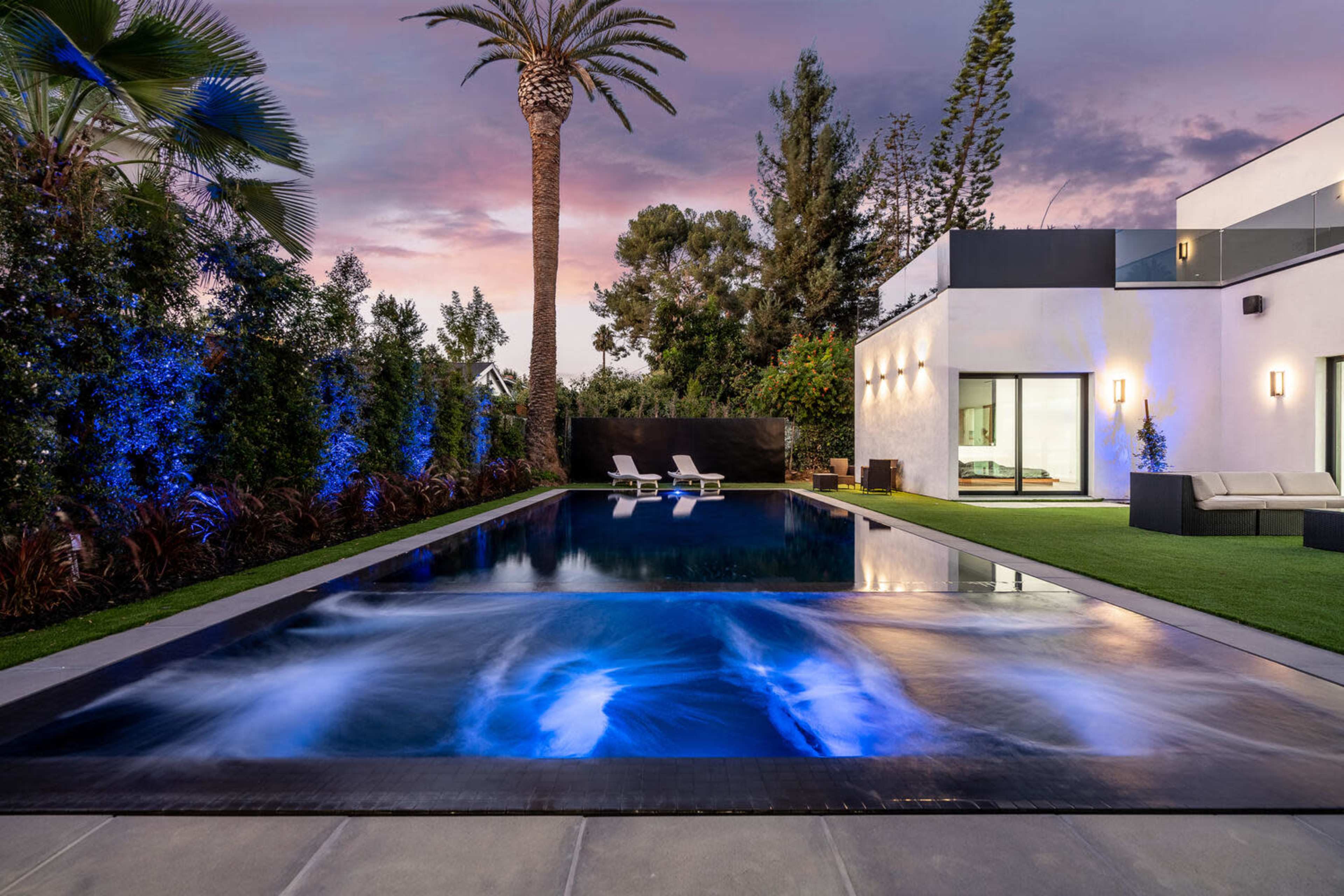 A modern backyard features a rectangular swimming pool with illuminated water, surrounded by lush greenery and palm trees, and a contemporary house in the background.