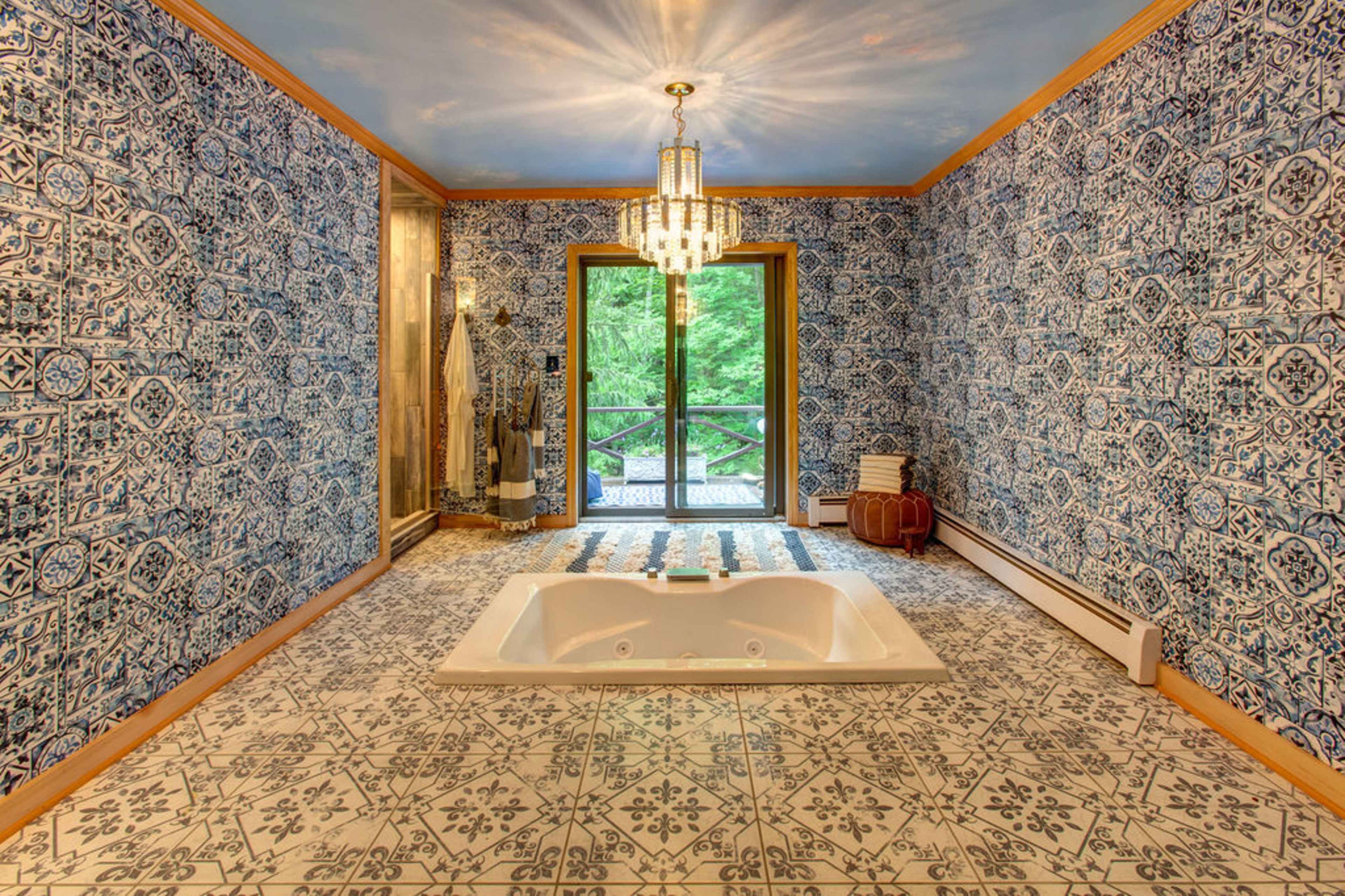A bathroom with blue patterned wallpaper, a white Jacuzzi tub, and a large window overlooking greenery.