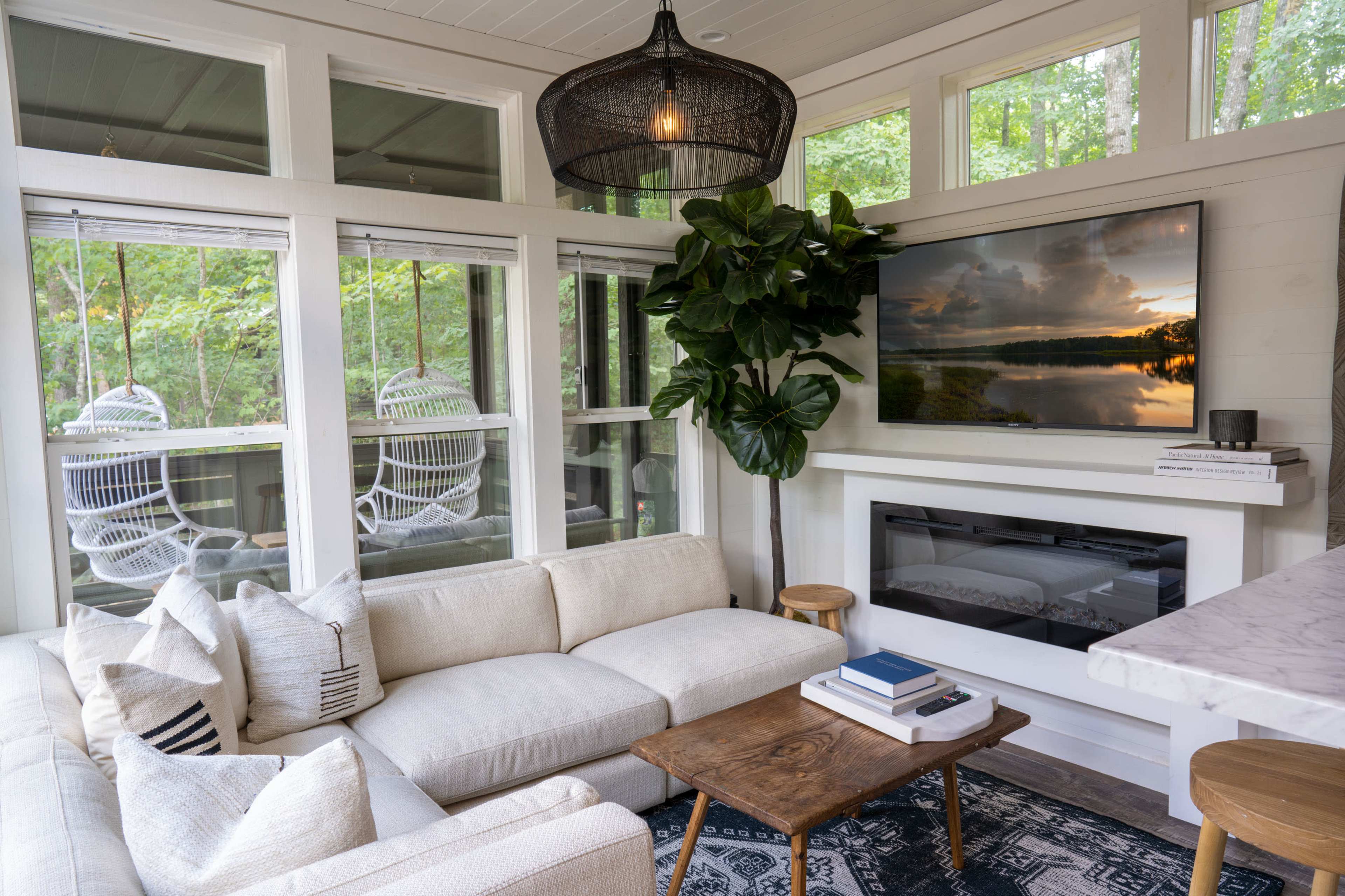 A cozy living room features a white sectional sofa, a wooden coffee table, and a mounted TV above a sleek fireplace, surrounded by large windows and greenery.
