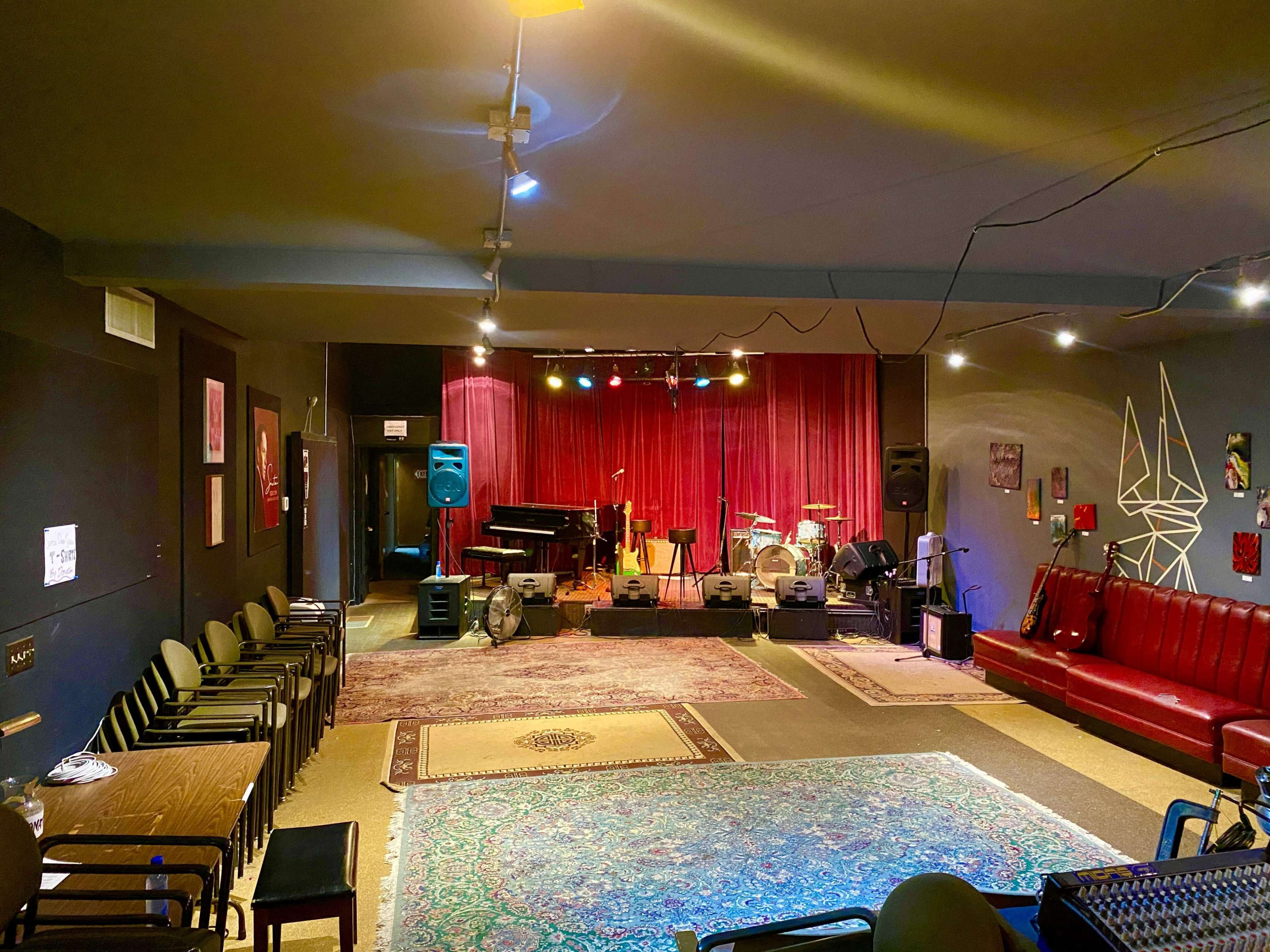 A music venue with a stage, various instruments set up, and seating arranged in front of a red curtain.