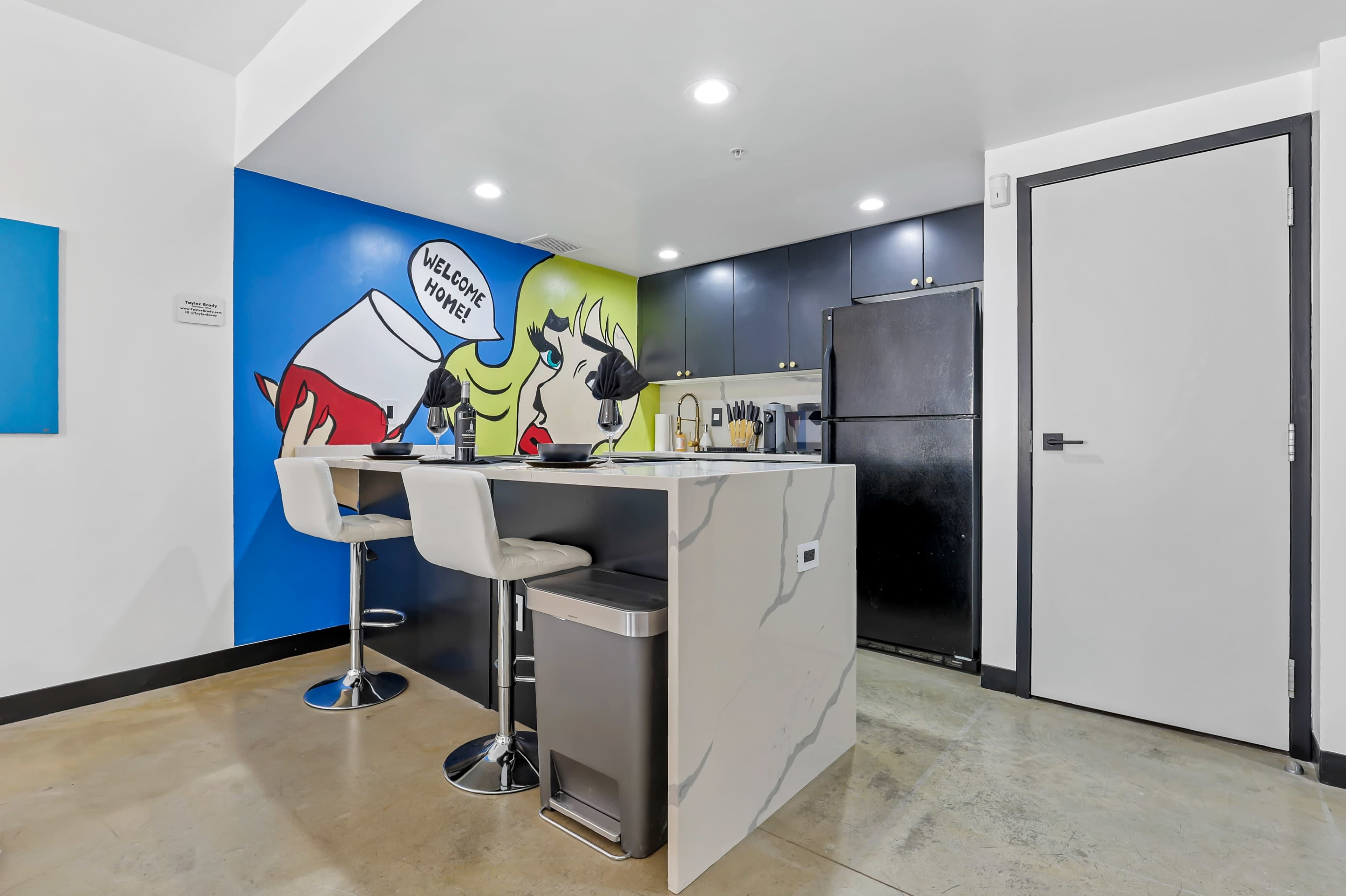 A modern kitchen features a bar with two stools and a colorful wall mural, welcoming guests.