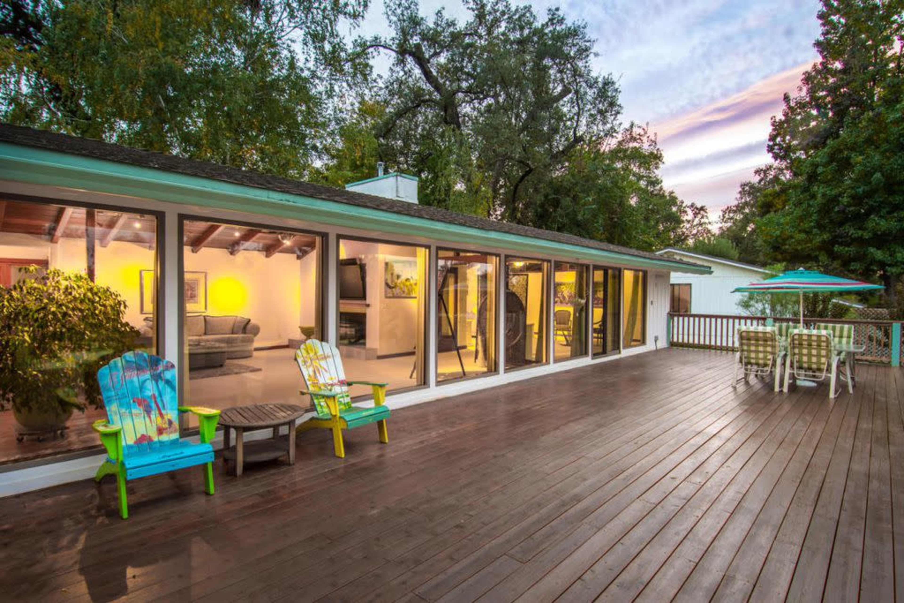 The image shows a modern house with large glass windows overlooking a wooden deck that features colorful outdoor furniture and a dining area.