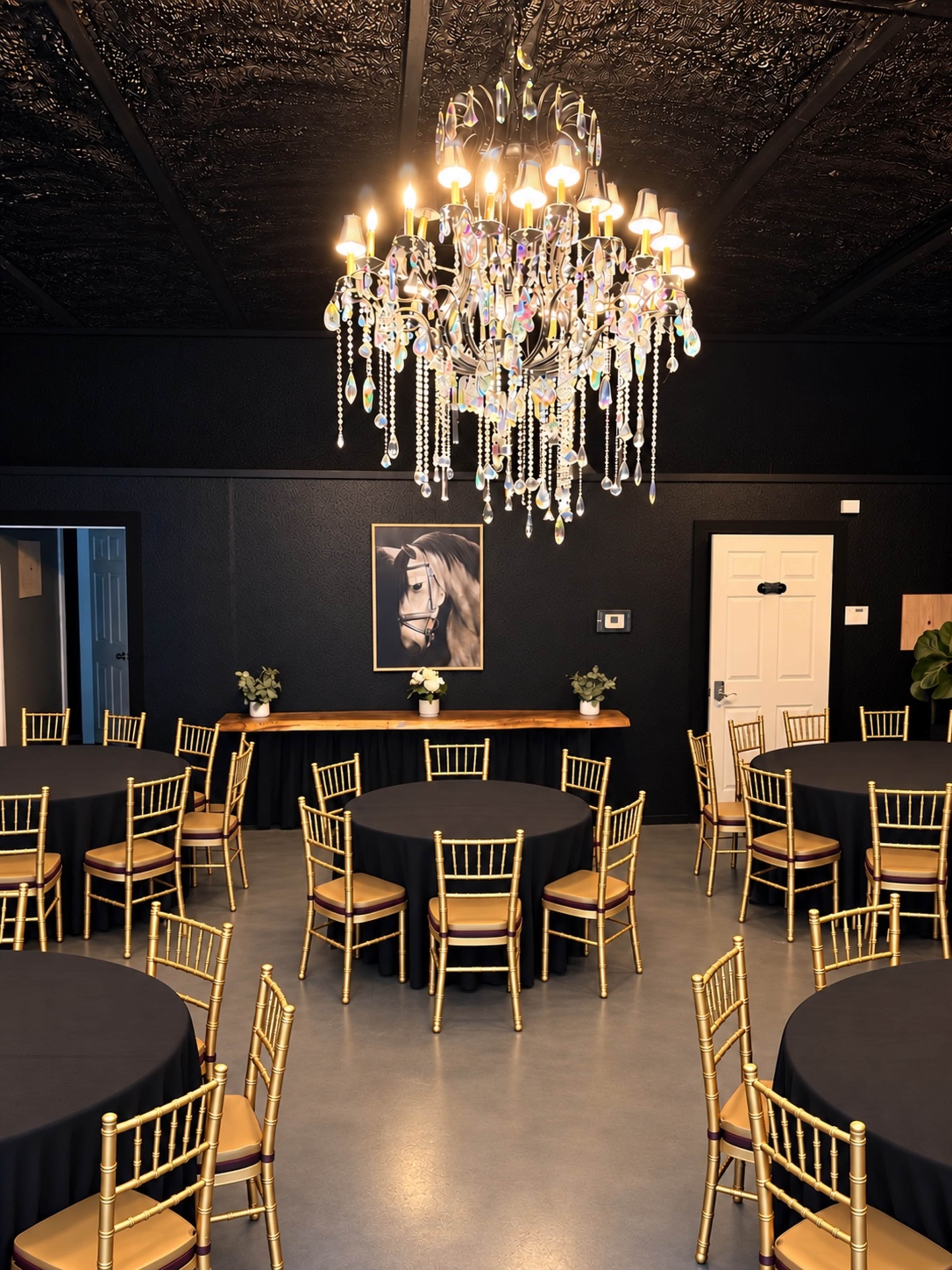 The image shows a lunchroom setup with several round tables covered in black tablecloths, golden chairs, a large chandelier overhead, and a framed portrait on the wall.