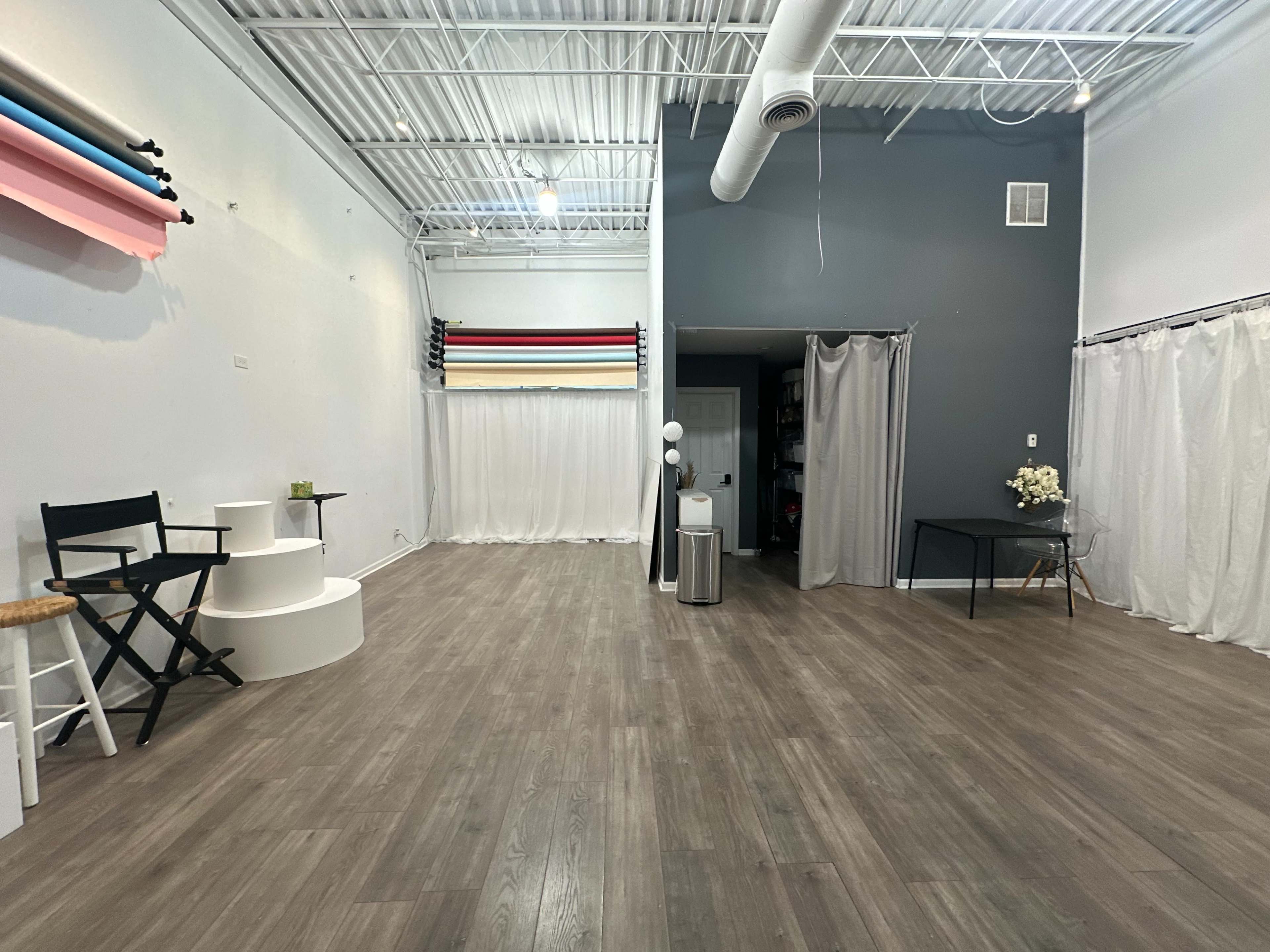 The image shows a spacious studio with wooden flooring, featuring a seating area, various backdrops on the wall, and draped curtains in the background.