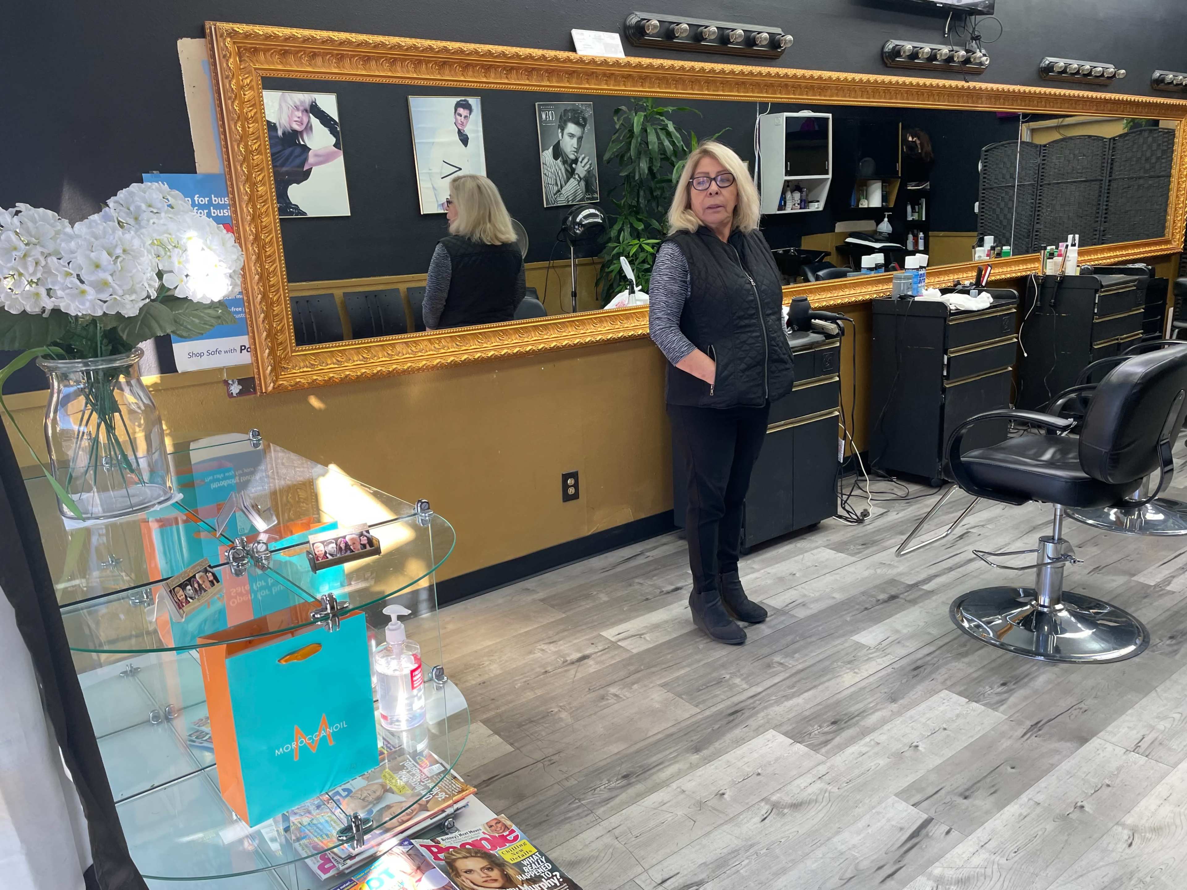 A woman stands in a hair salon beside a large mirror, with styling tools and magazines displayed on a glass table.
