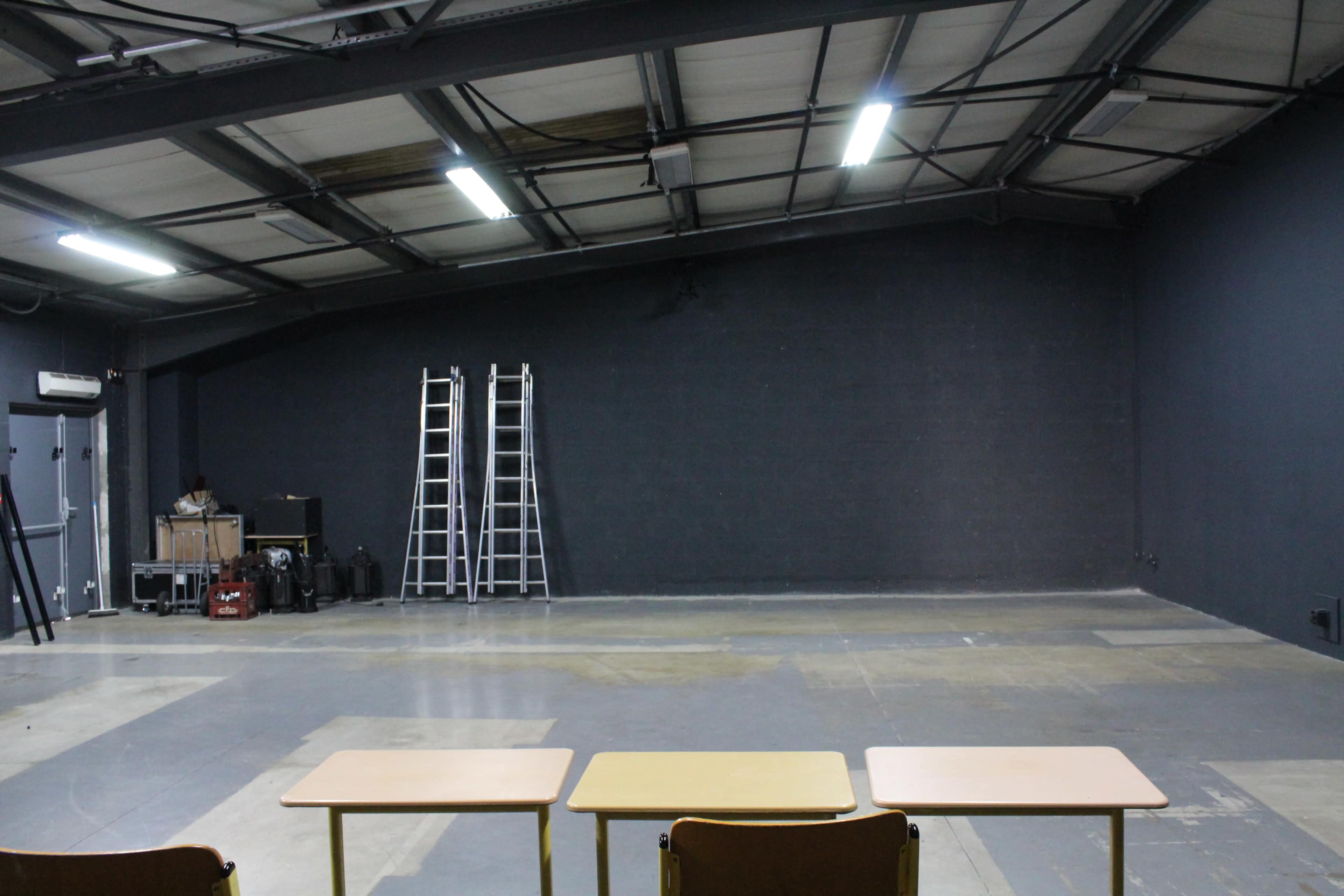 An empty industrial space with three tables in the foreground, two ladders leaning against a wall, and various equipment in the background.
