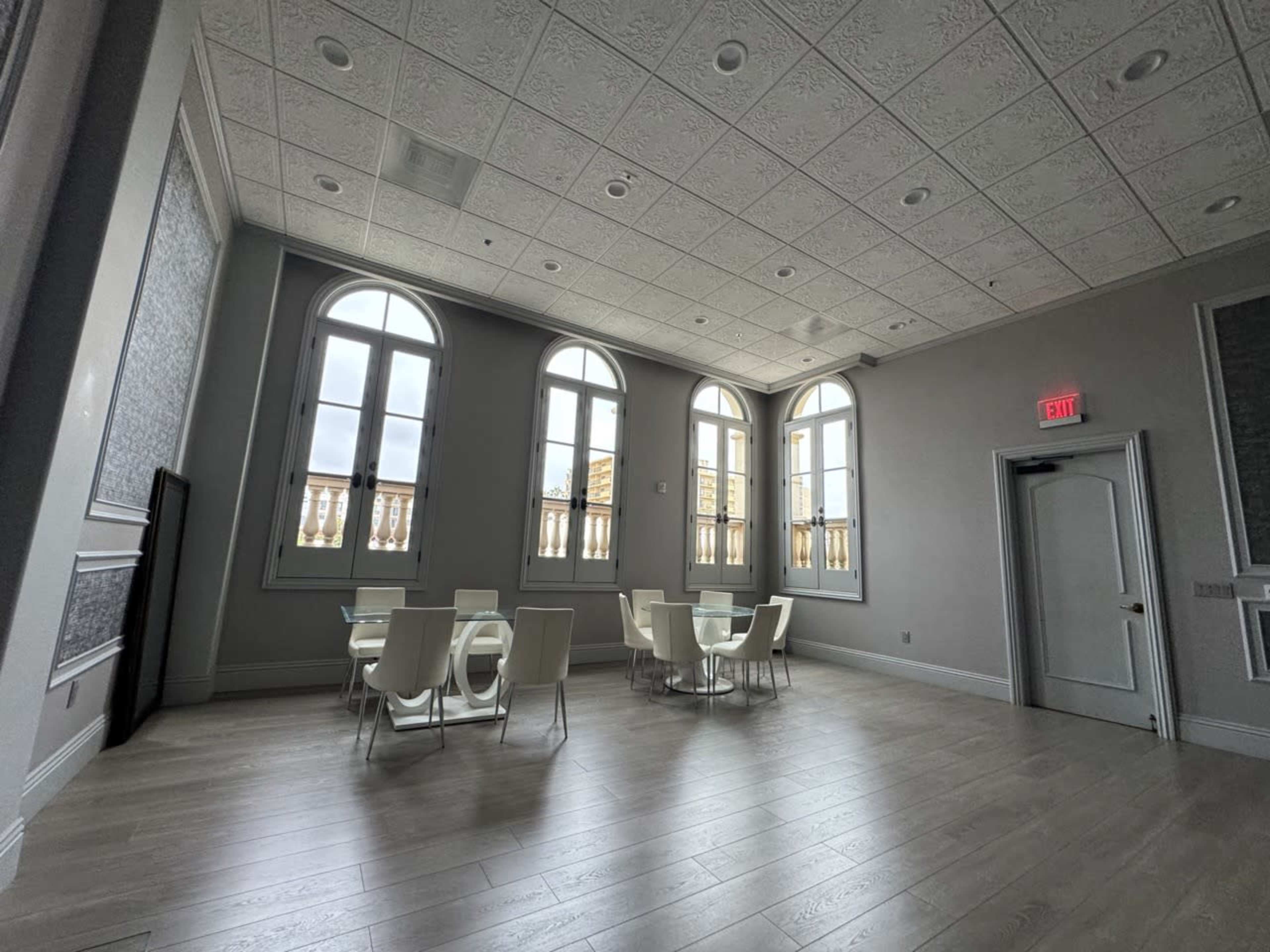 The image shows a spacious room with large arched windows, wooden flooring, and modern white furniture, including tables and chairs.