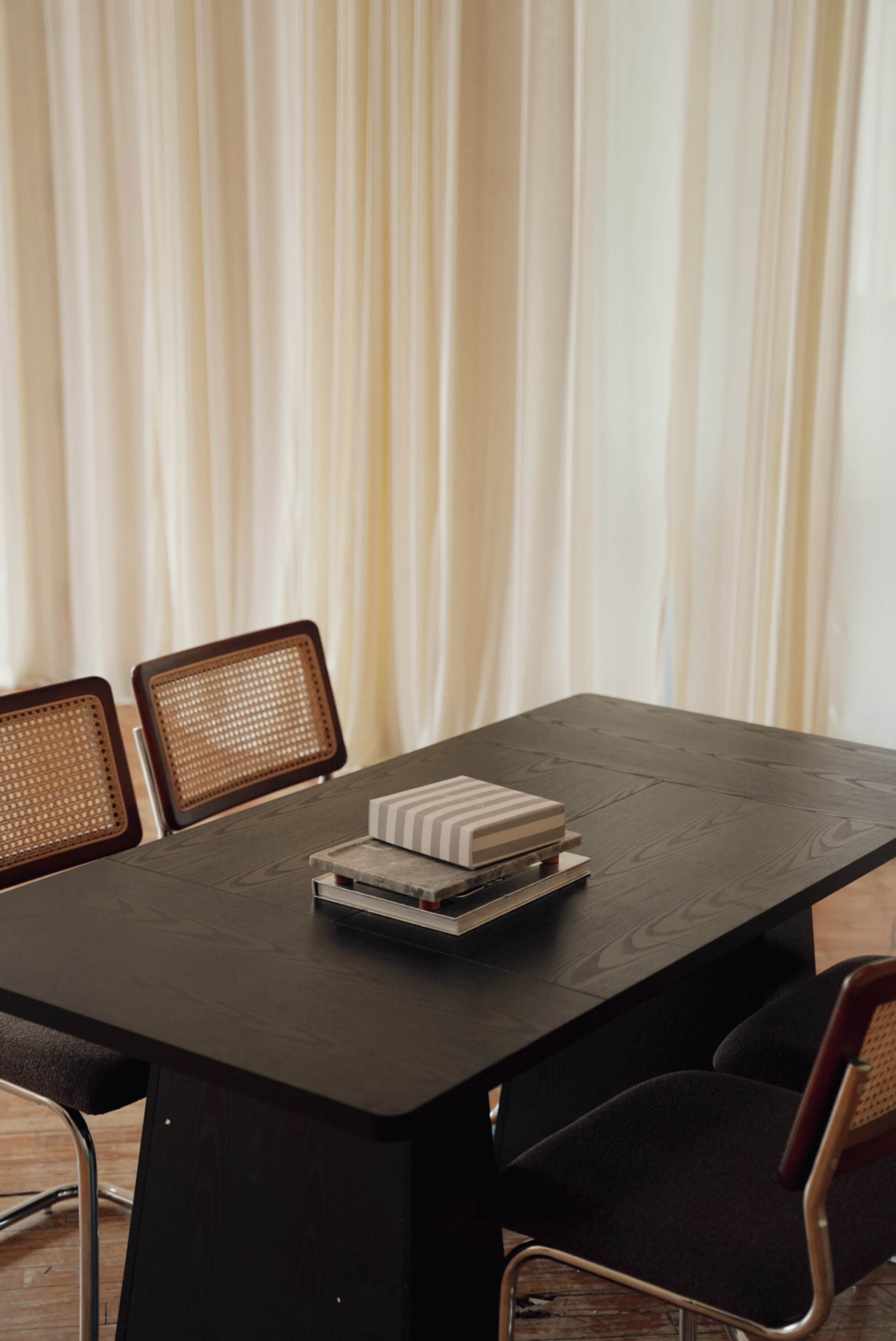 A dark wooden table with a stack of books and a decorative item is surrounded by four cane-back chairs in a room with light draped curtains.