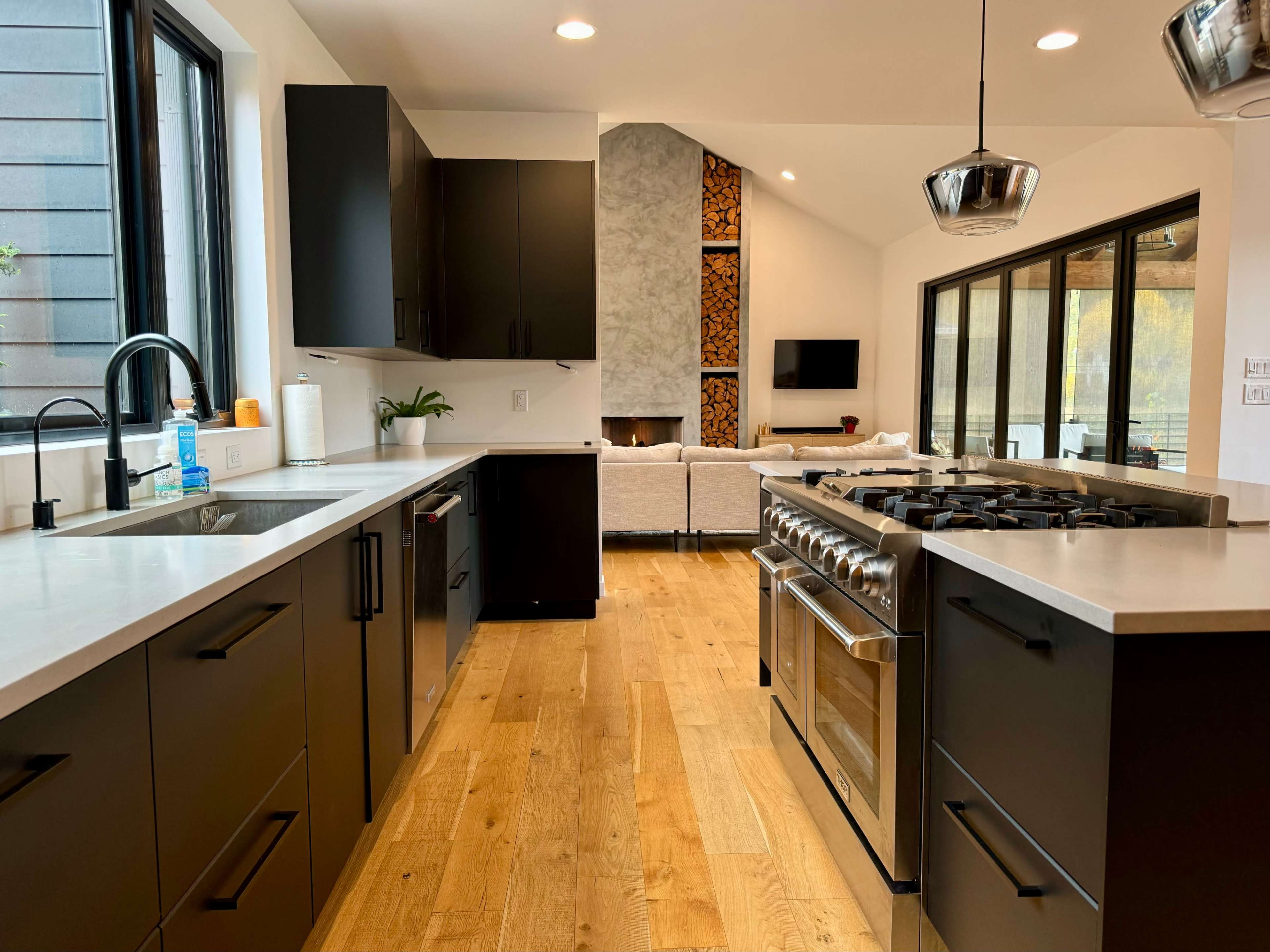 A modern kitchen featuring black cabinets, a stainless steel stove, and an open layout leading to a living area with a fireplace.