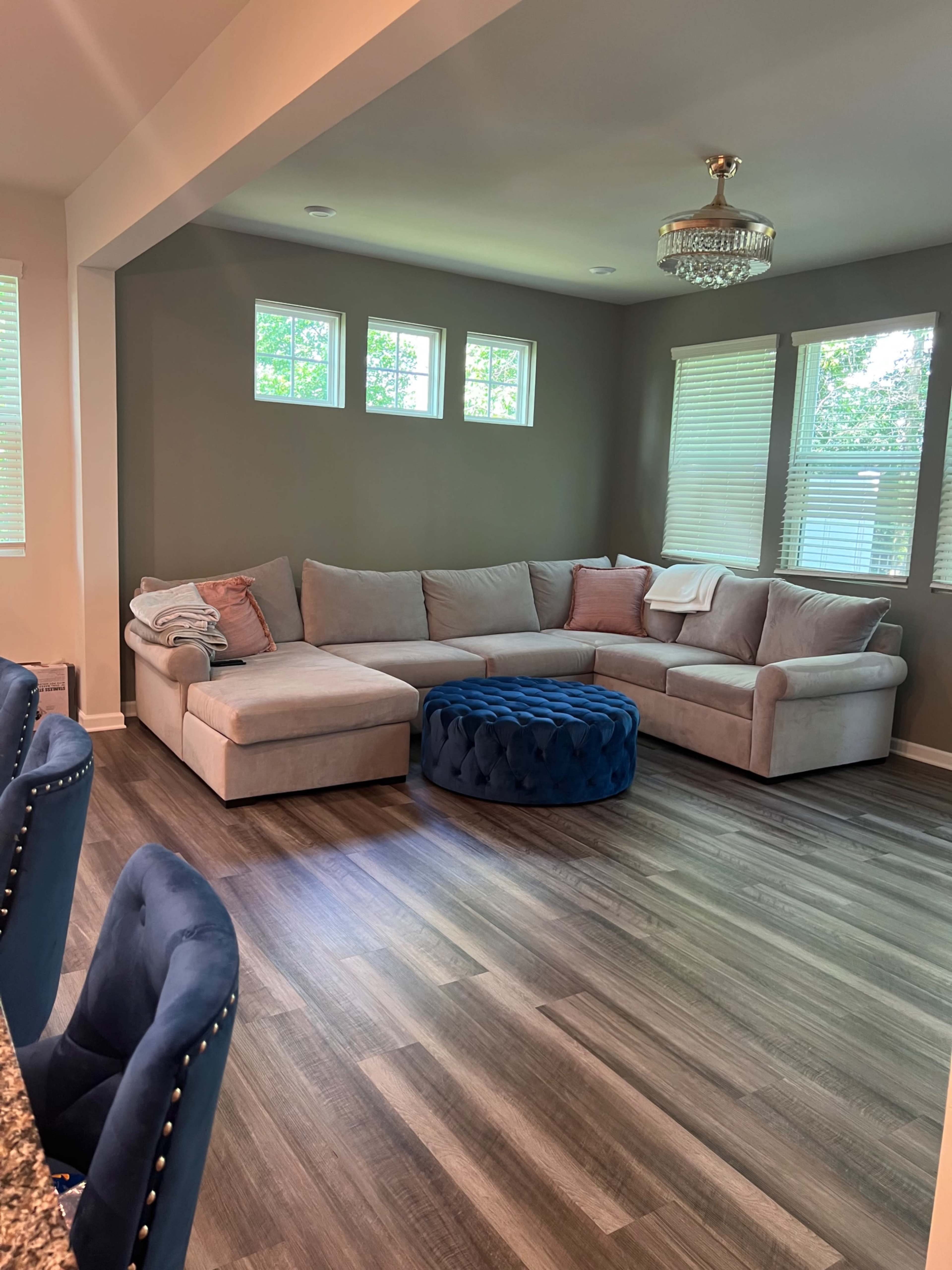 A living room with a light-colored sectional sofa, a round blue ottoman, and several windows allowing natural light.