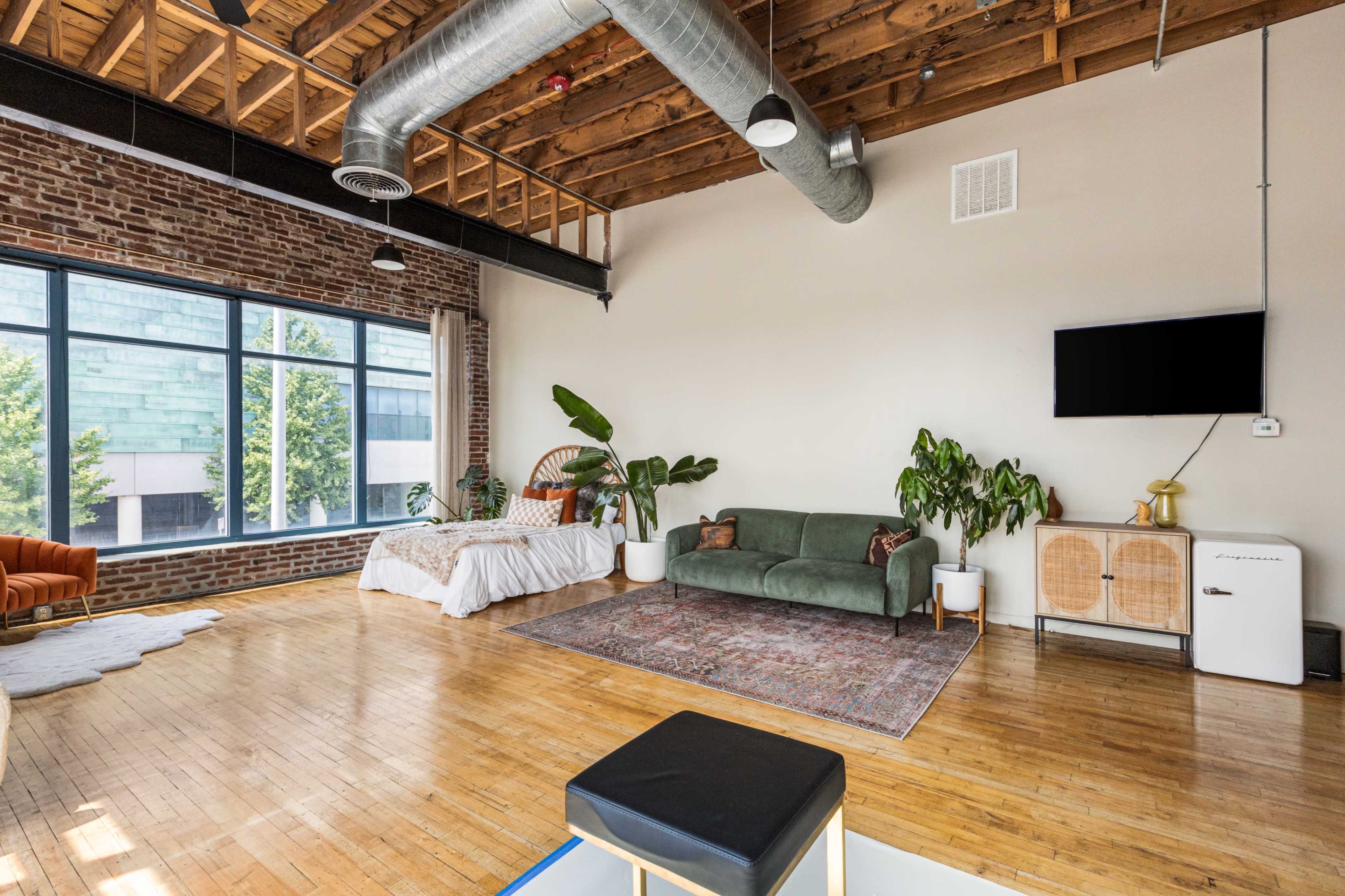 The image depicts a spacious, airy living area featuring a bed, a green sofa, potted plants, and a large window with a view.