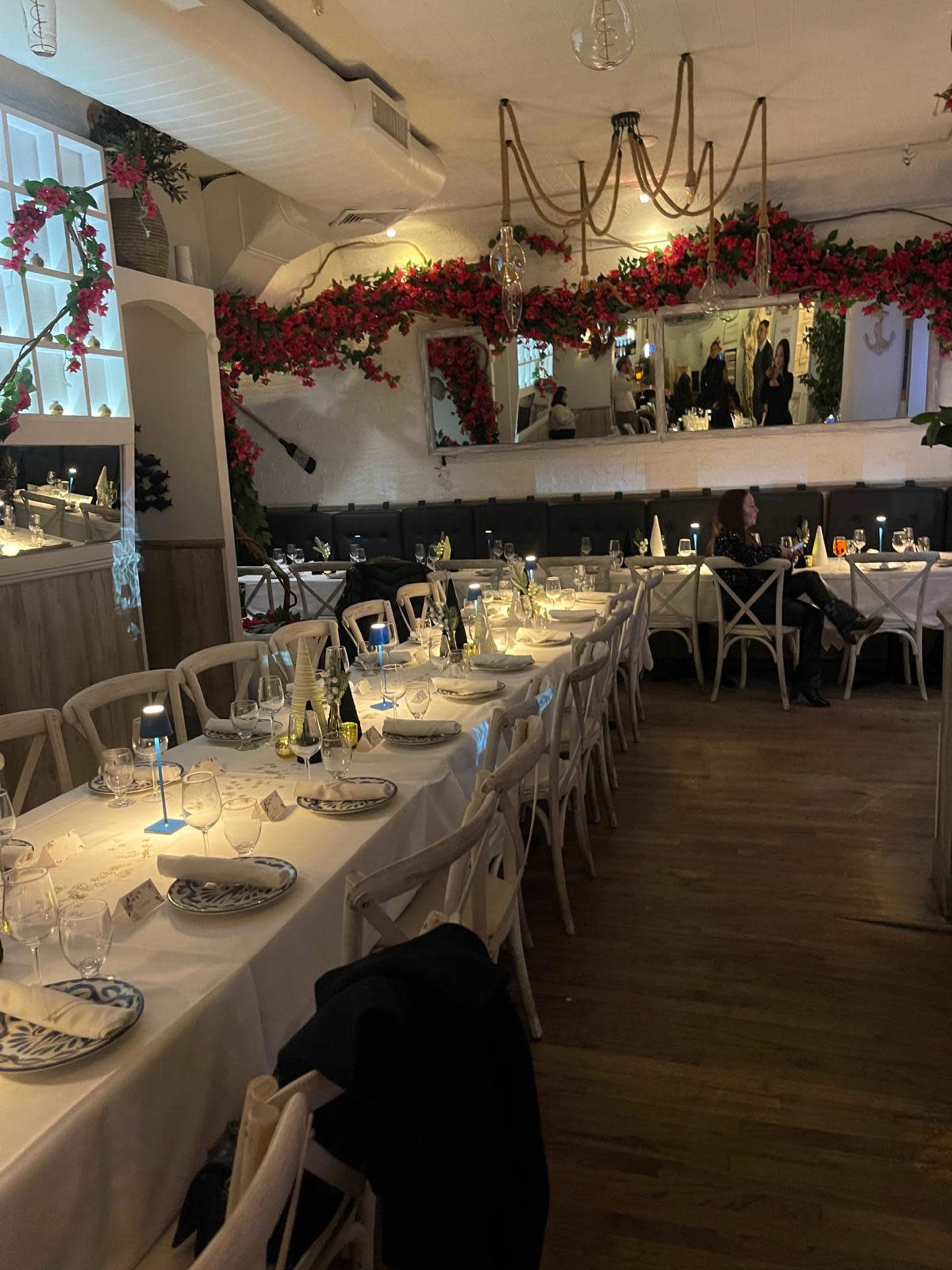 The image shows a restaurant interior with long tables set for dining, adorned with white tablecloths and elegant place settings, surrounded by floral decorations along the walls.