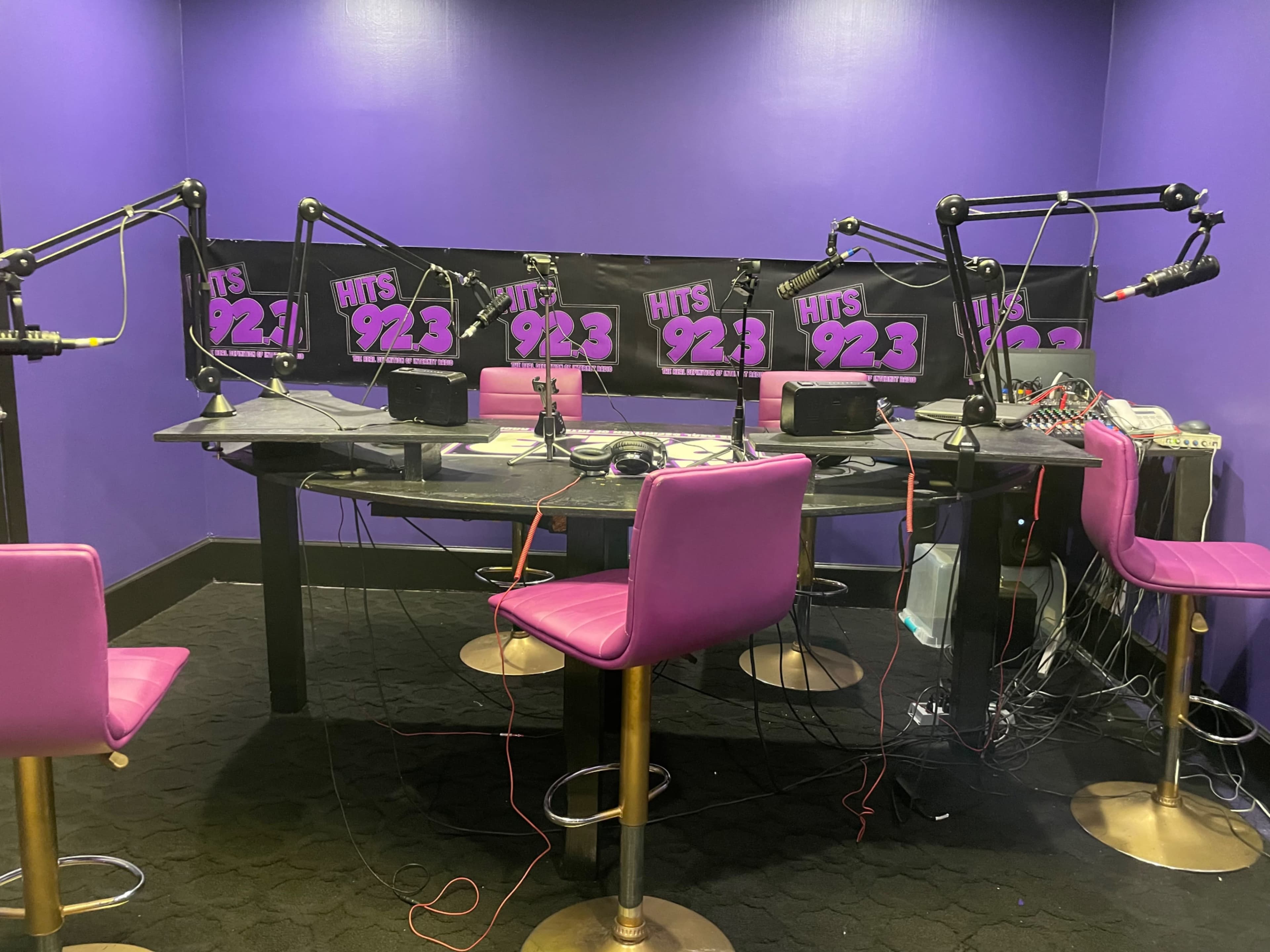 A radio studio with a purple wall, a circular table equipped with microphones, and several pink stools arranged around it.