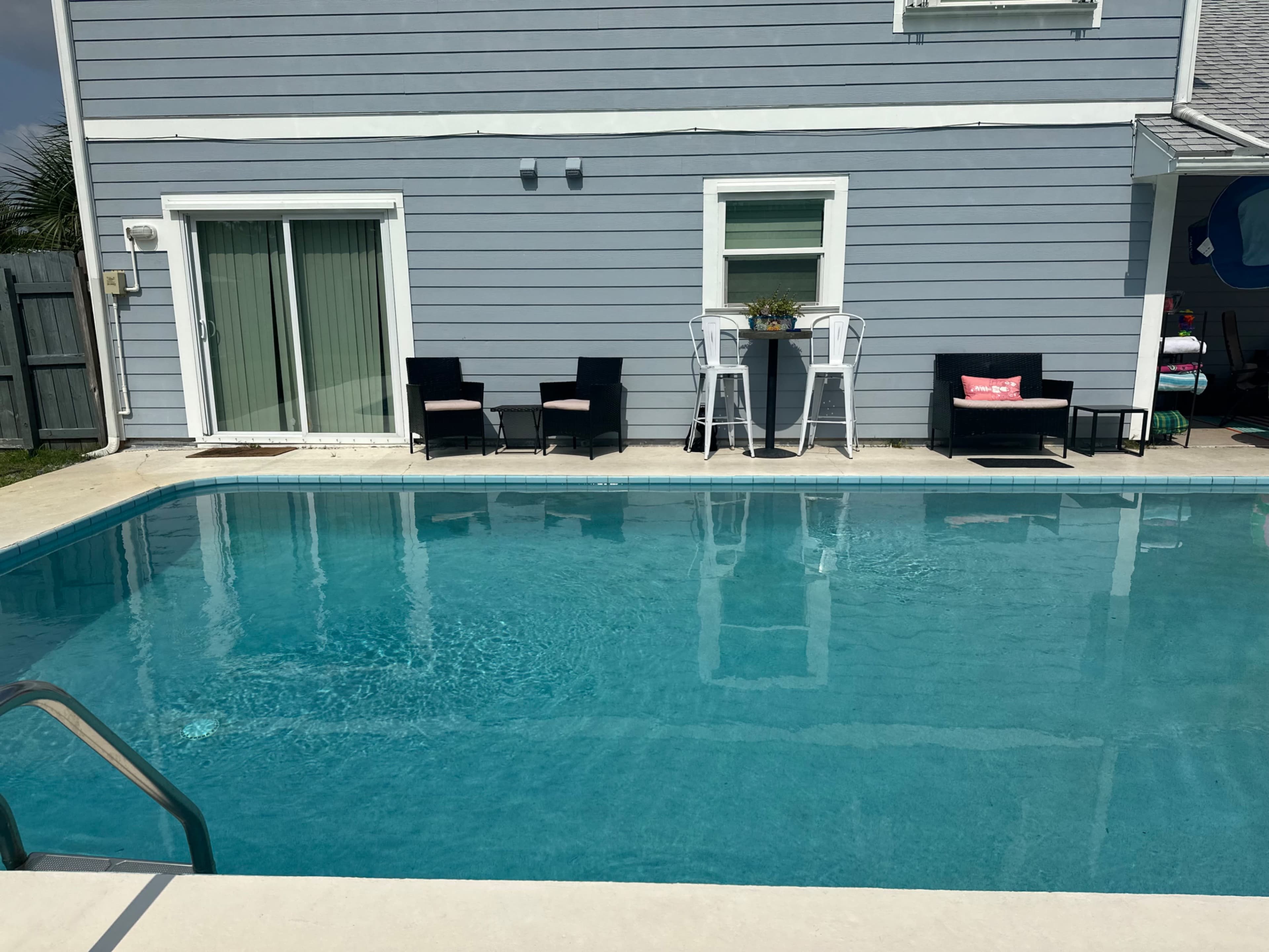 The scene features a clear swimming pool in front of a light blue house with outdoor seating and a small table.