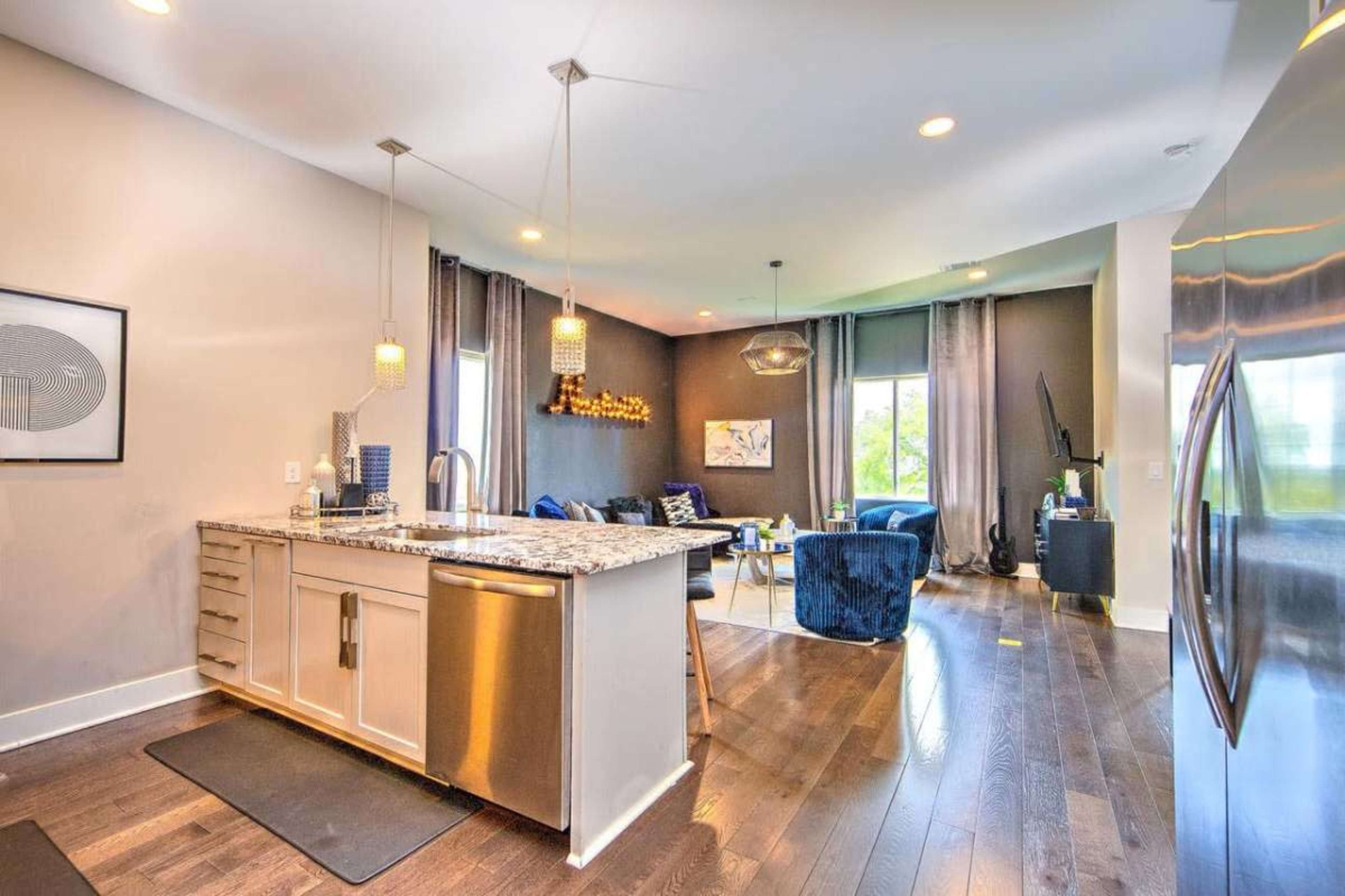 The image shows a modern kitchen with a granite countertop, stainless steel appliances, and a view into a living area featuring blue furniture and decorative lighting.