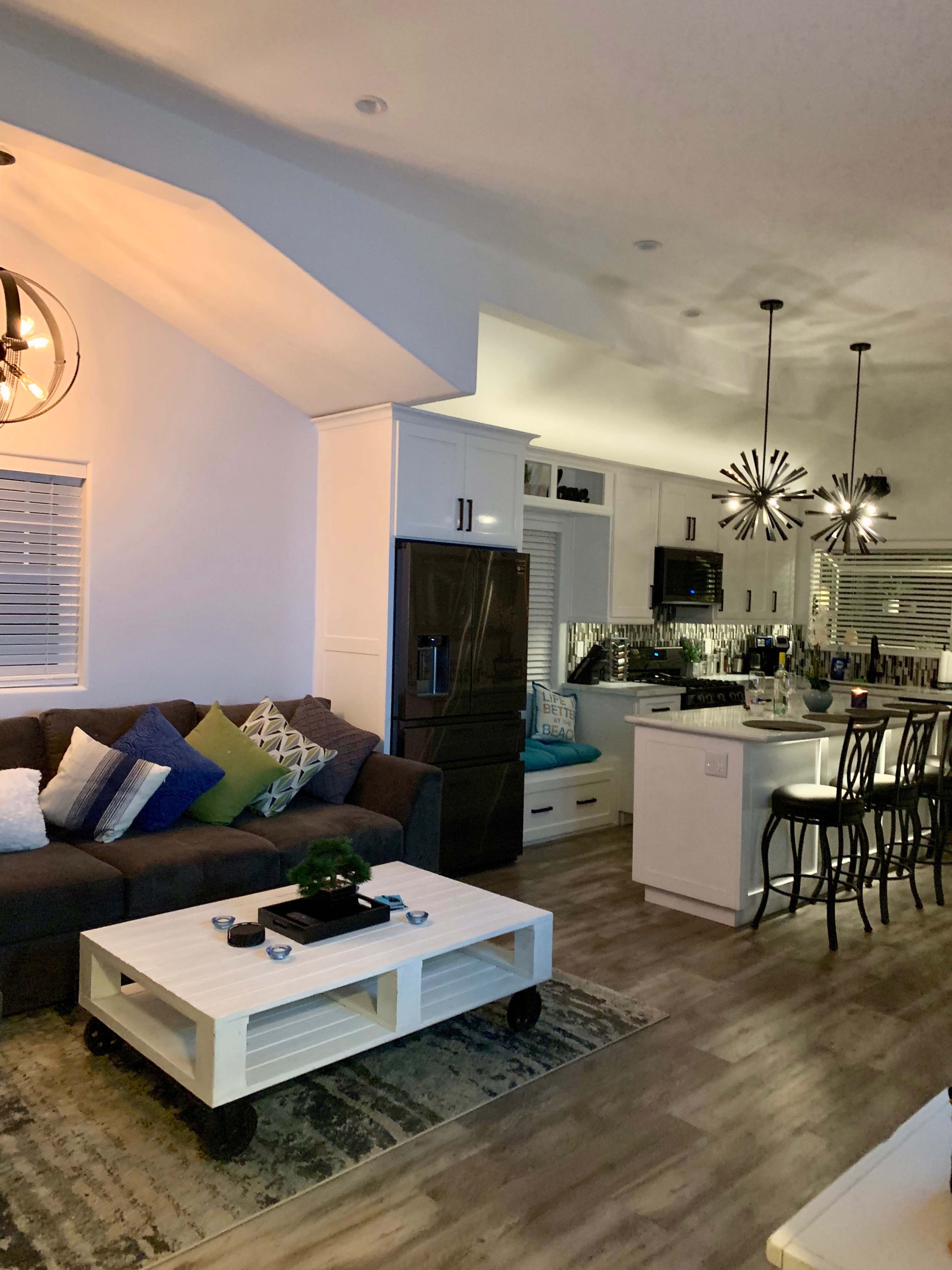 The image shows a modern living room and kitchen area with a sectional sofa, a white coffee table, and pendant lighting above the kitchen island.