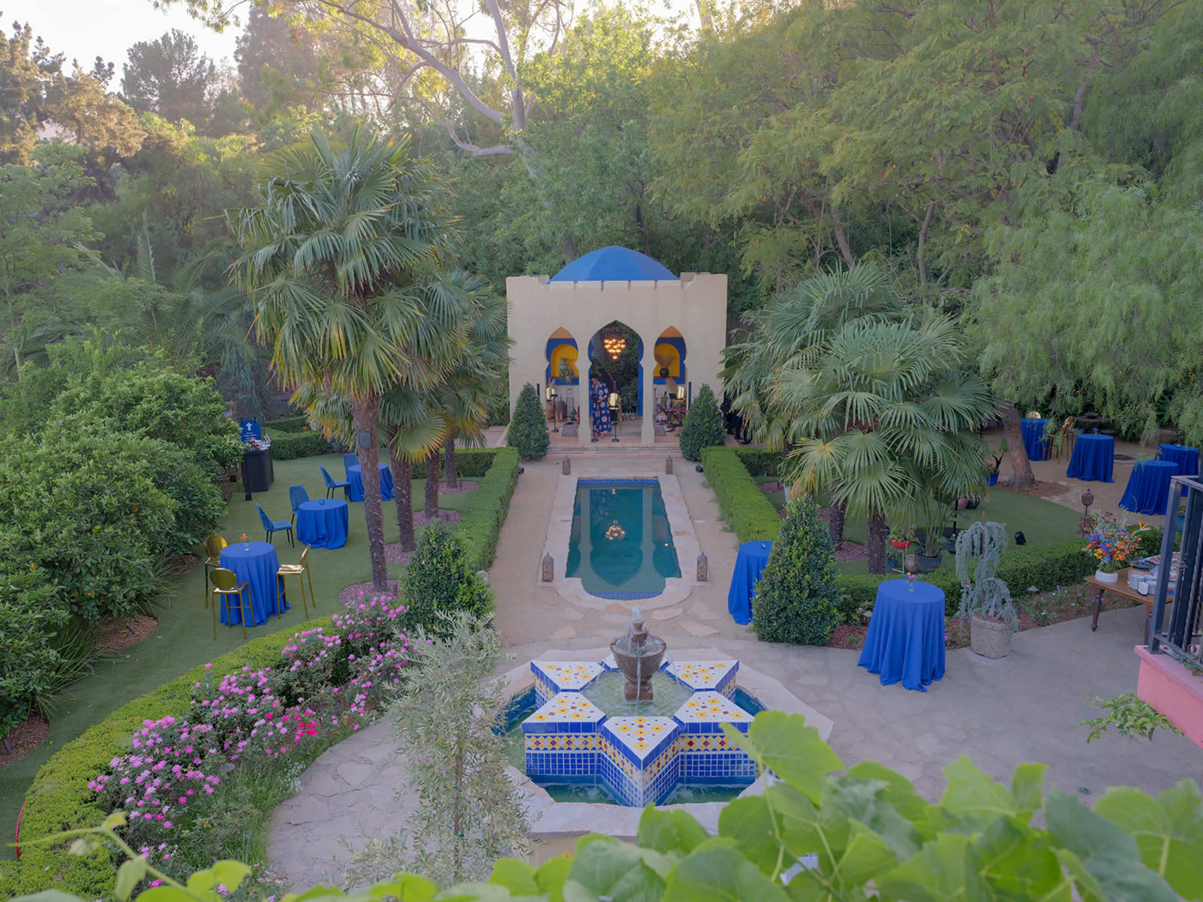 The image shows a landscaped outdoor garden with a central pool, a decorative fountain, and surrounded by lush greenery and tables set for an event.