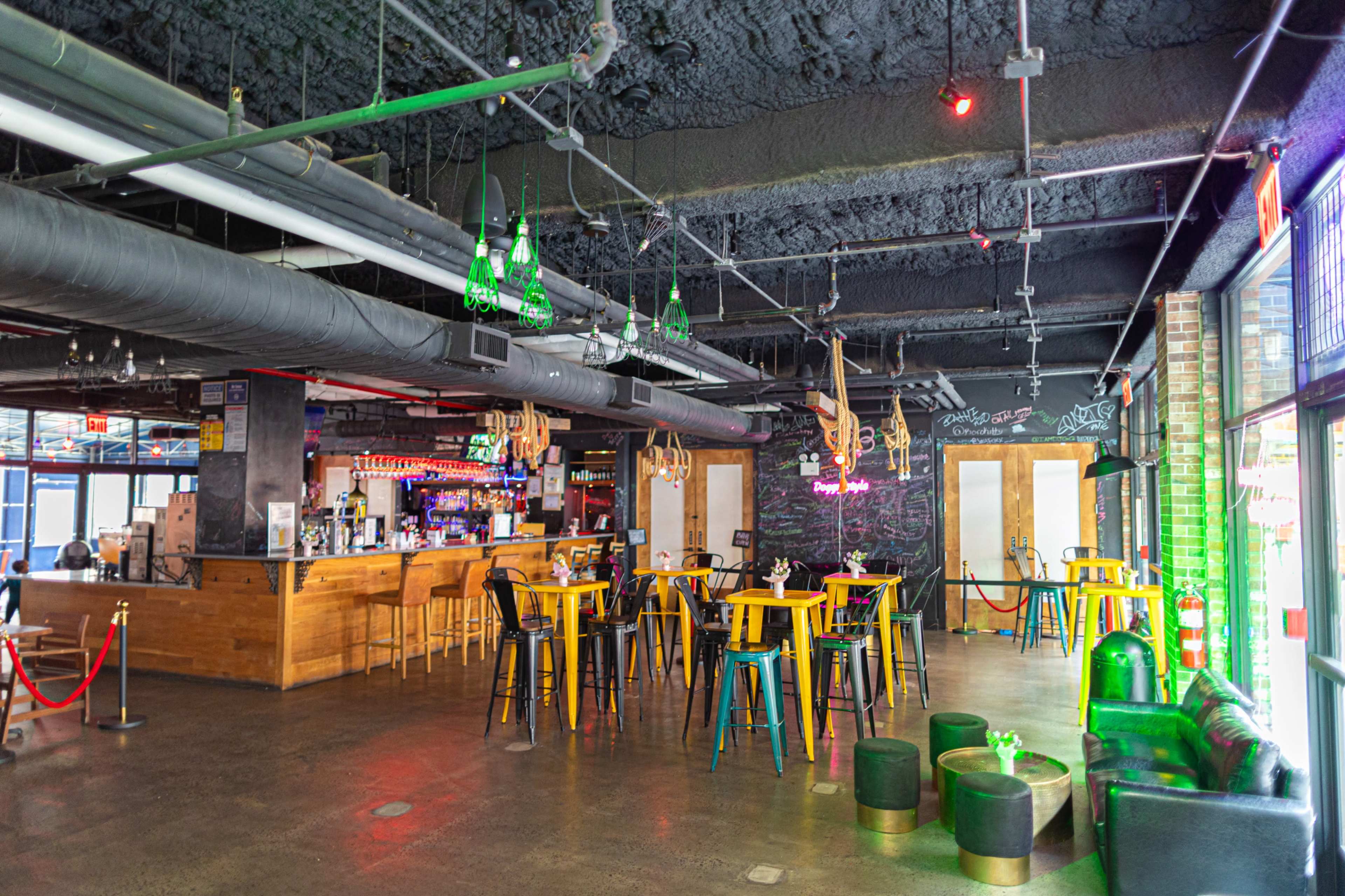 The image shows a modern bar interior with a wooden counter, colorful tables and stools, and a graffiti-decorated wall.