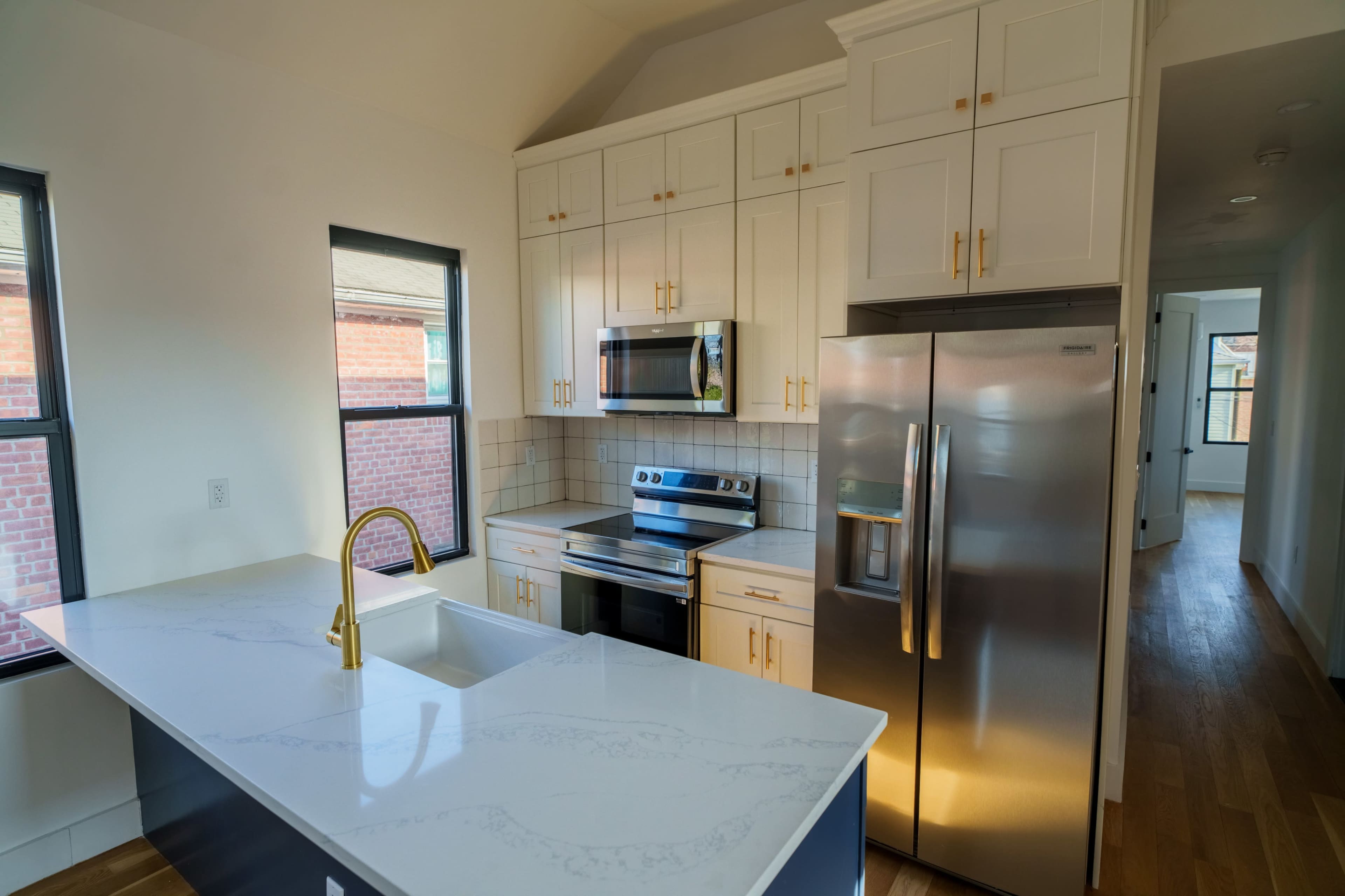 The image shows a modern kitchen with white cabinets, a stainless steel refrigerator, and a large island with a sink and a gold faucet.