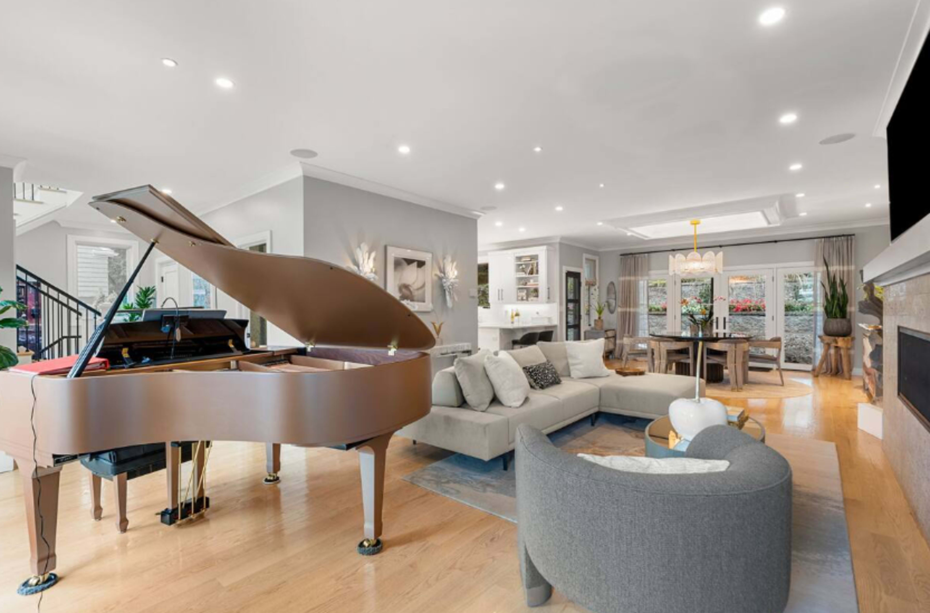 A grand piano is positioned in a modern living room that features a sectional sofa, a dining area, and a large window overlooking an outdoor space.