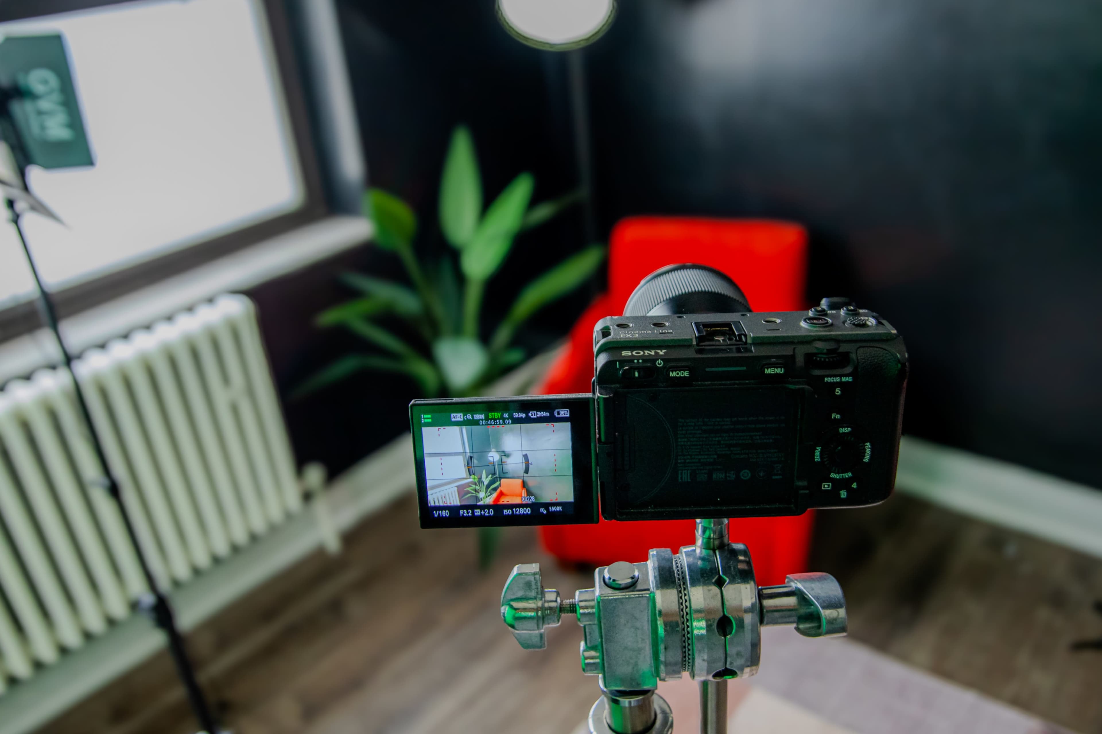 The image shows a camera positioned on a tripod facing a sitting area with a red chair and a potted plant in a room with dark walls.
