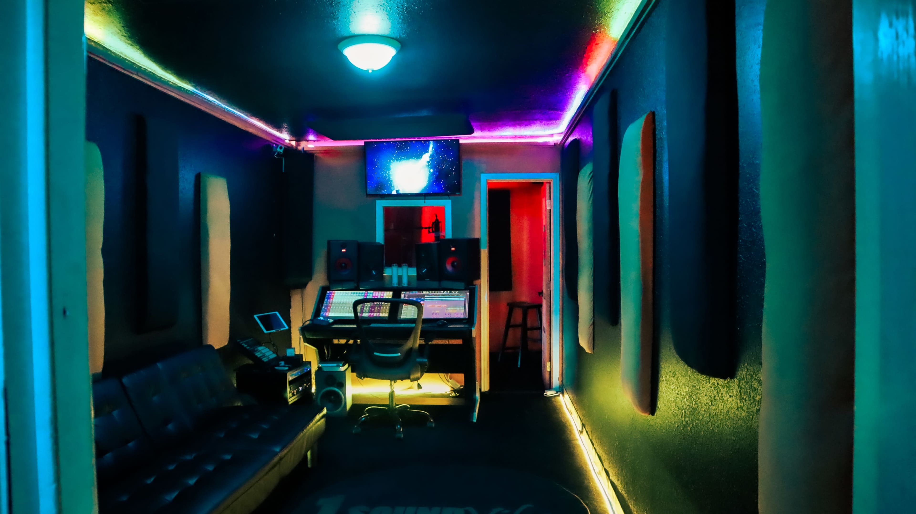A music recording studio features a mixing desk, a computer monitor, and colorful ambient lighting against dark walls.