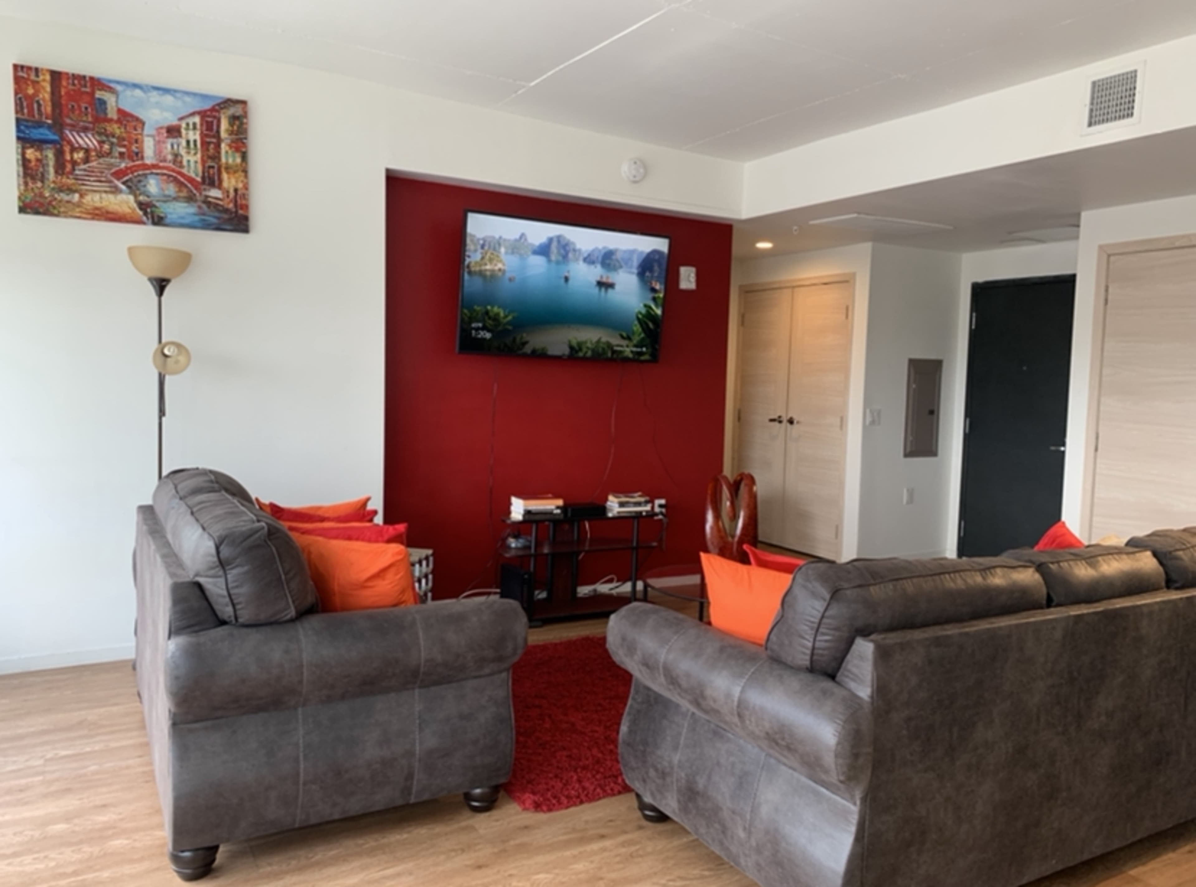 The image shows a living room with two gray sofas facing a wall-mounted television, a red accent wall, and a piece of artwork depicting a scenic view.