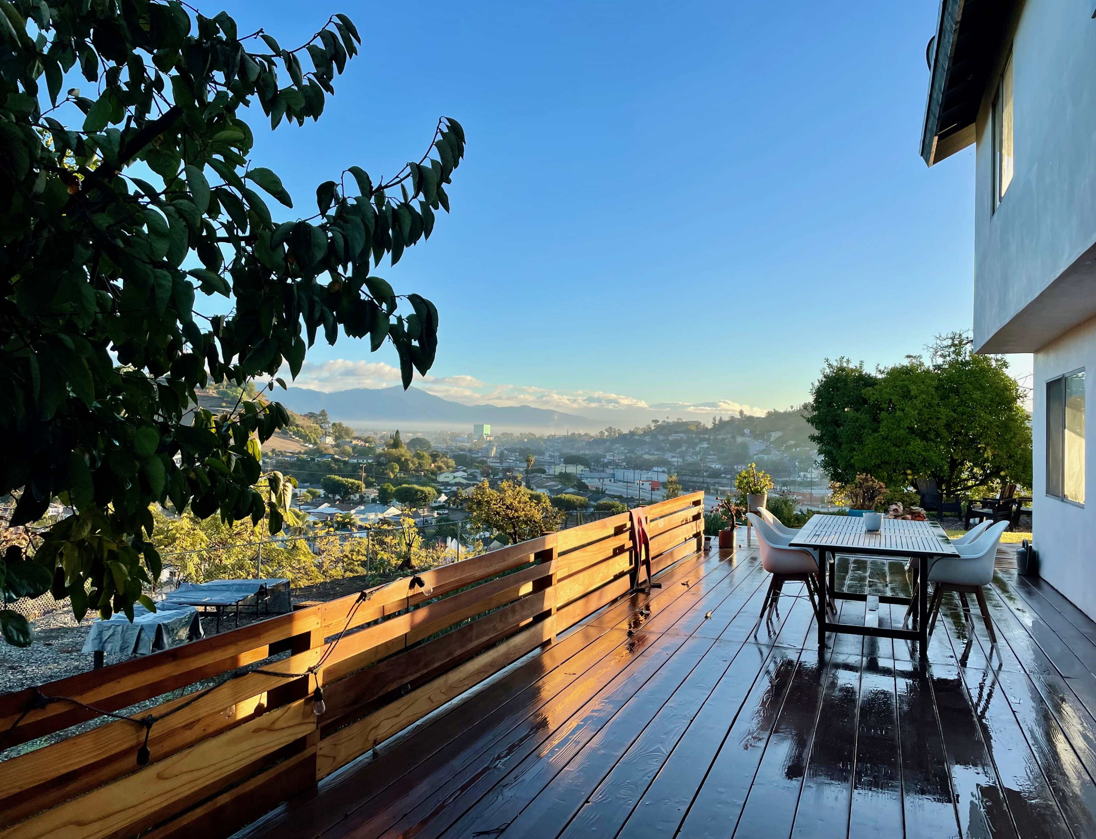 The image shows a wooden deck with a table and chairs overlooking a scenic view of a town and distant hills under a clear blue sky.