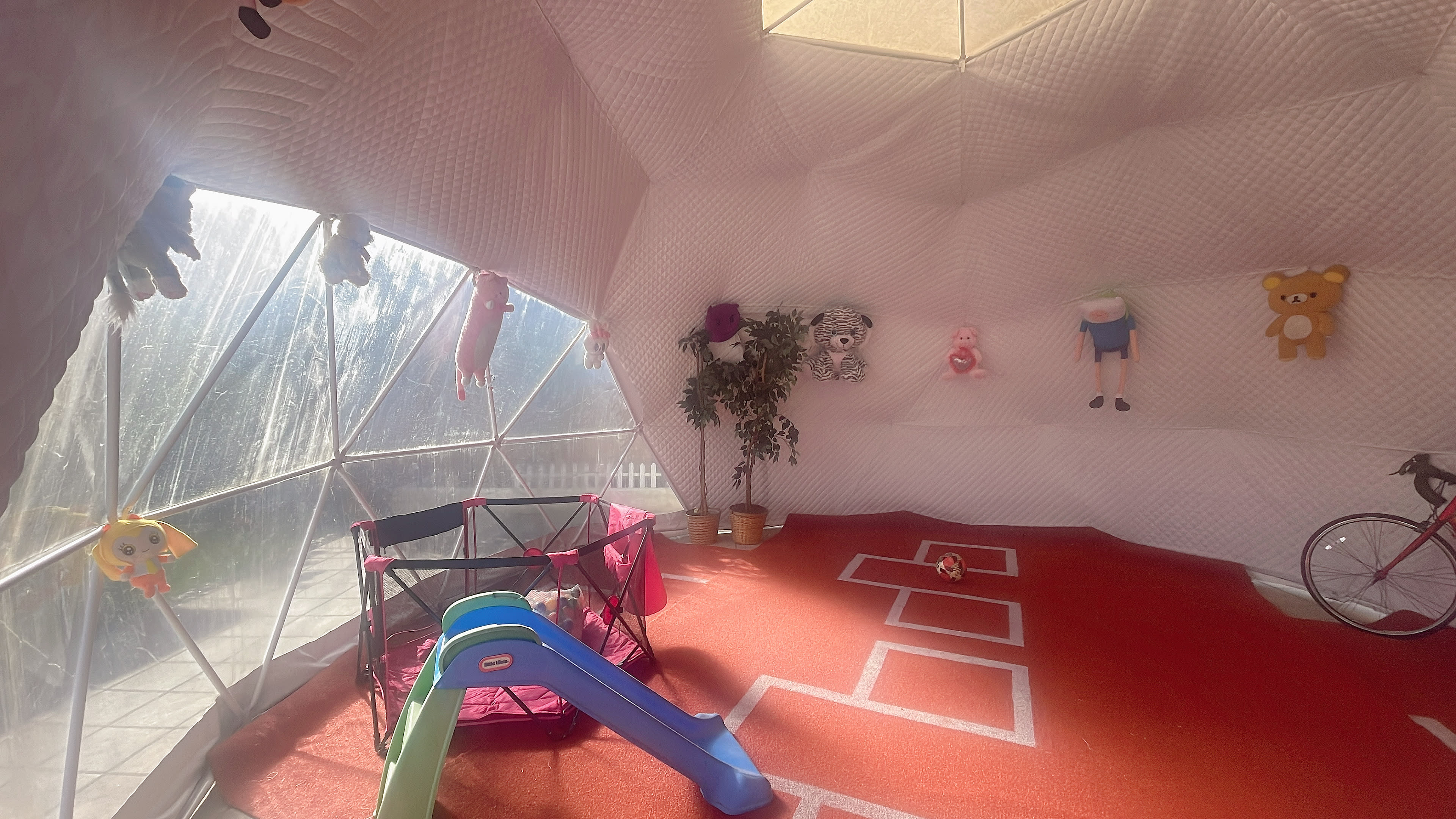 The interior of a geodesic dome features colorful stuffed toys hanging on the walls, a small slide, and a playpen on a red carpeted floor.