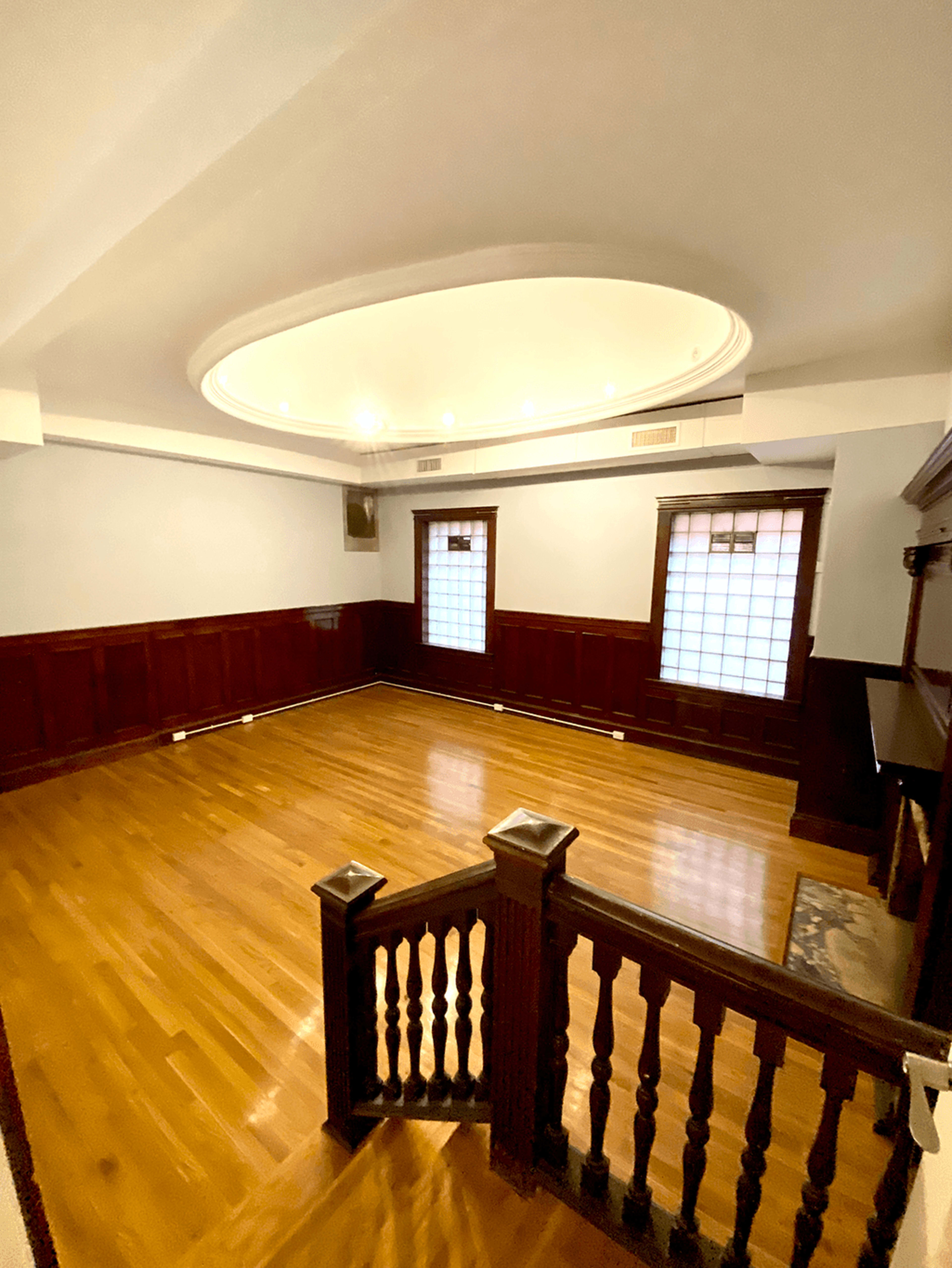 An empty room with wooden flooring, circular ceiling lights, and wainscoted walls, featuring two windows with grid patterns.