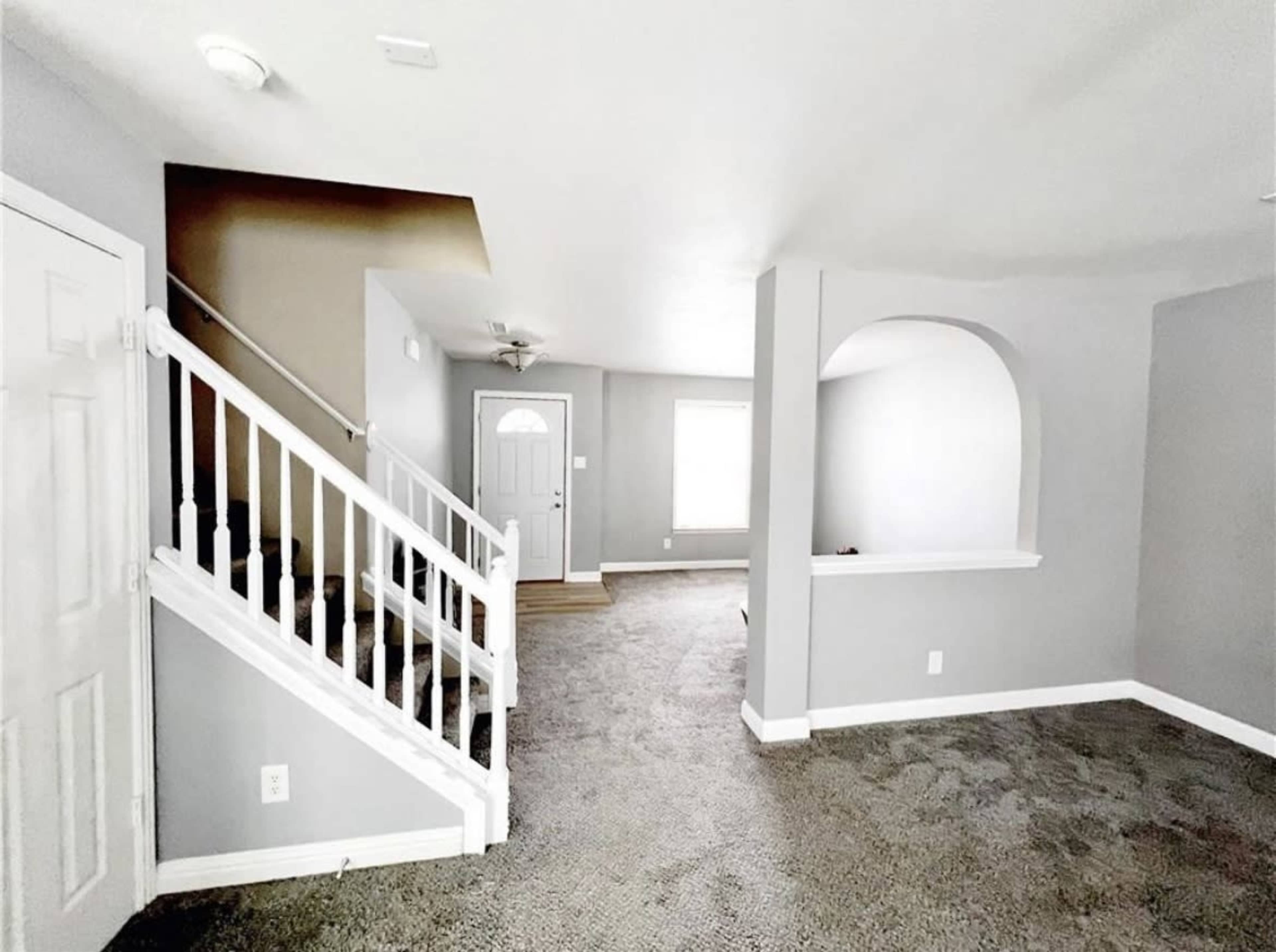 A gray-toned living room features a staircase, an arched entryway, and a front door leading to natural light.