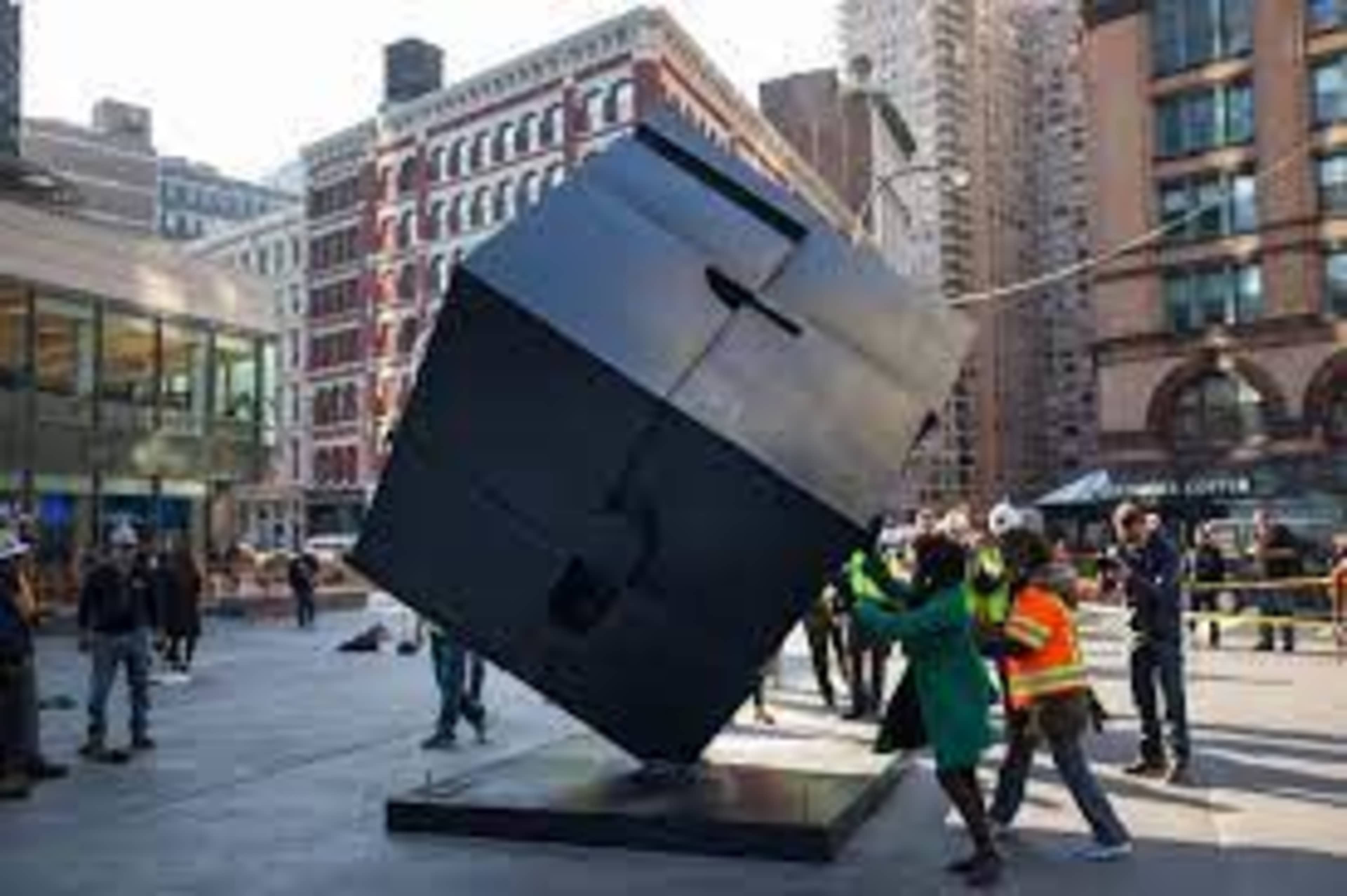 Workers in safety vests maneuver a large black cube sculpture in a city plaza surrounded by buildings.