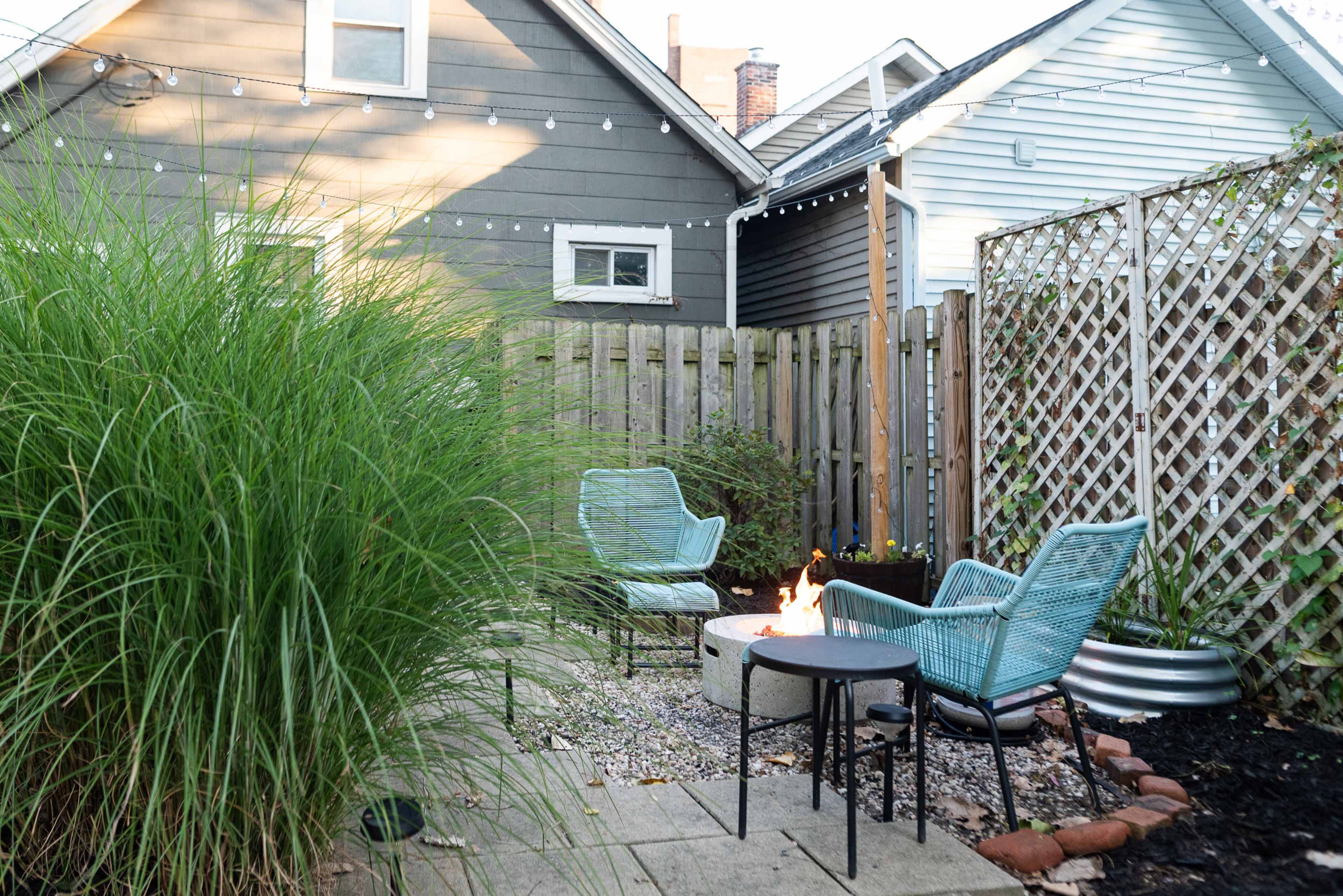 A small outdoor patio features two blue chairs, a circular fire pit with a flame, and surrounding greenery and a wooden fence.