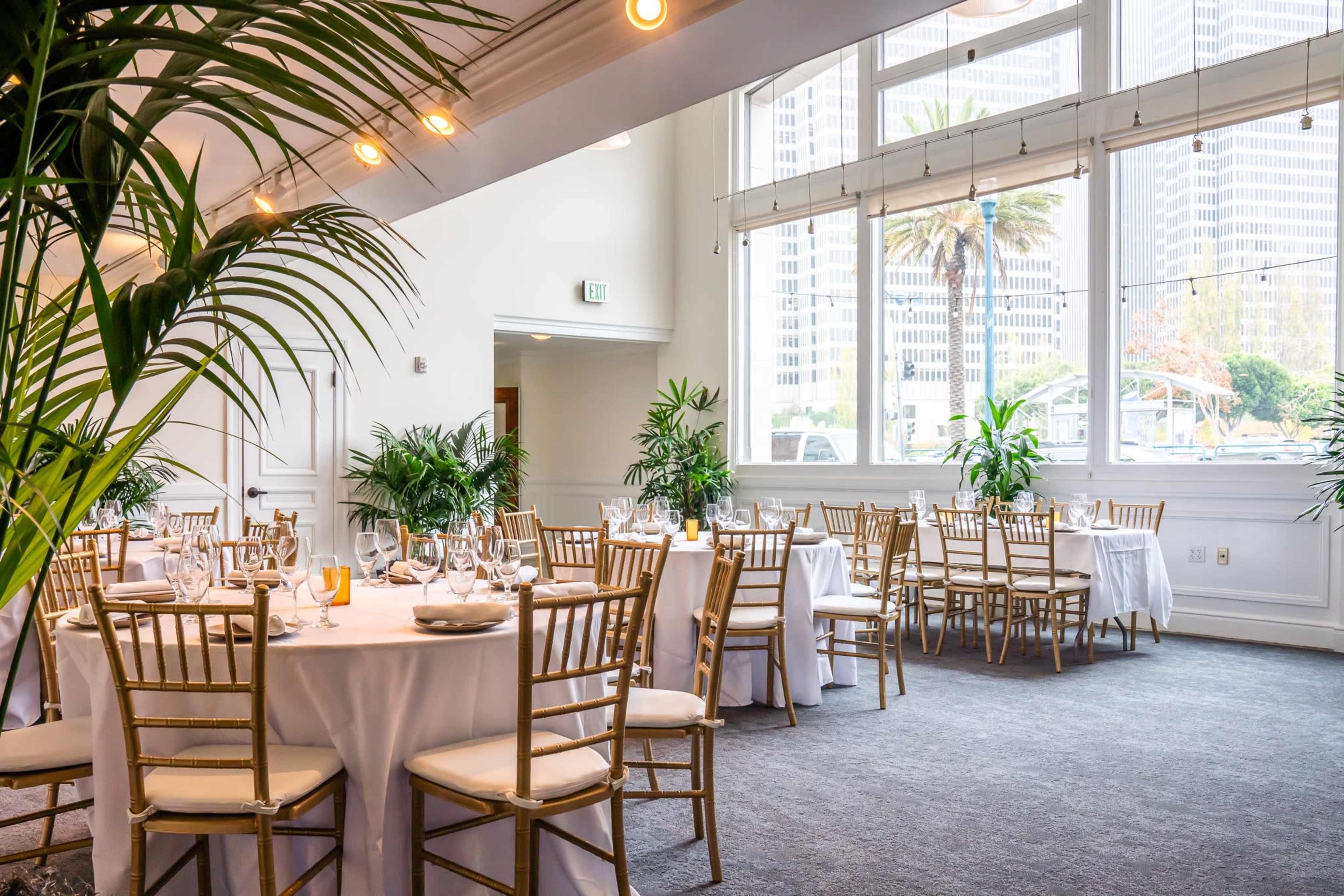 A banquet room with round tables dressed in white tablecloths and golden chairs, surrounded by large windows and indoor plants.