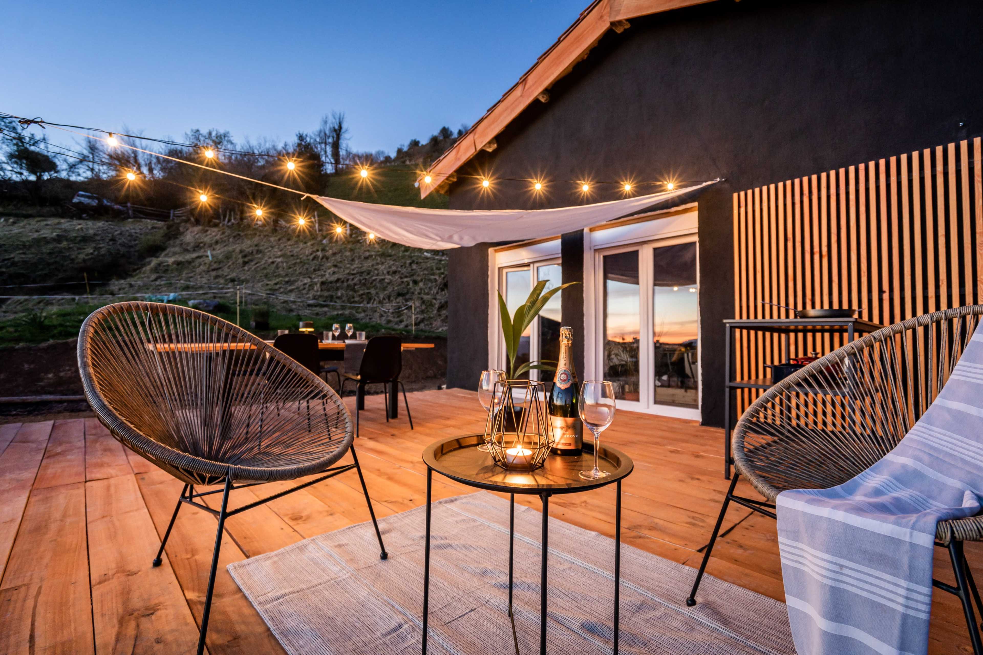 The image shows a cozy patio area with two chairs and a small table adorned with candles and a bottle of champagne, surrounded by softly glowing string lights against a backdrop of a house and a scenic landscape.