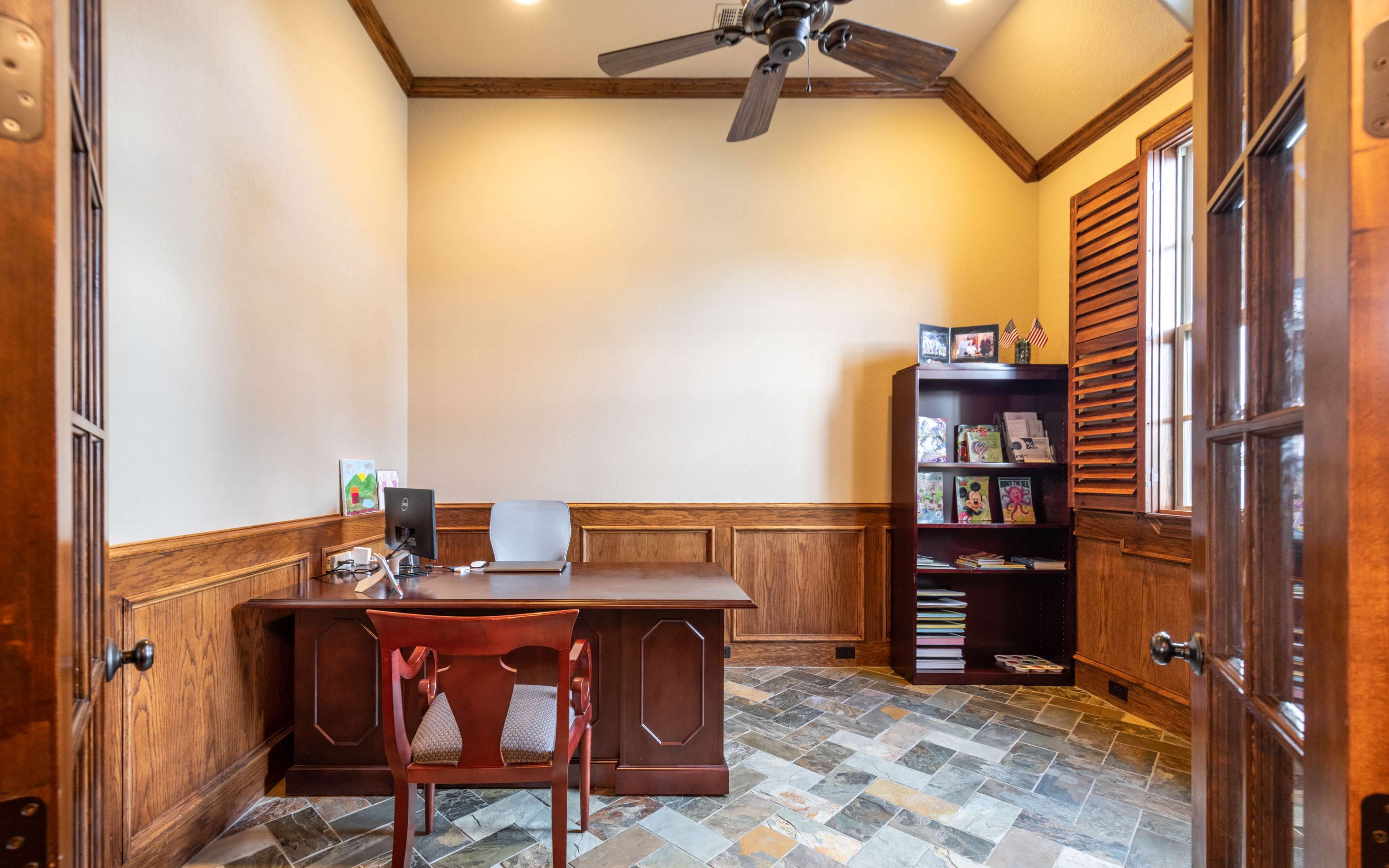 A wooden office with a desk, computer, and shelves filled with books, set against a slate tile floor.