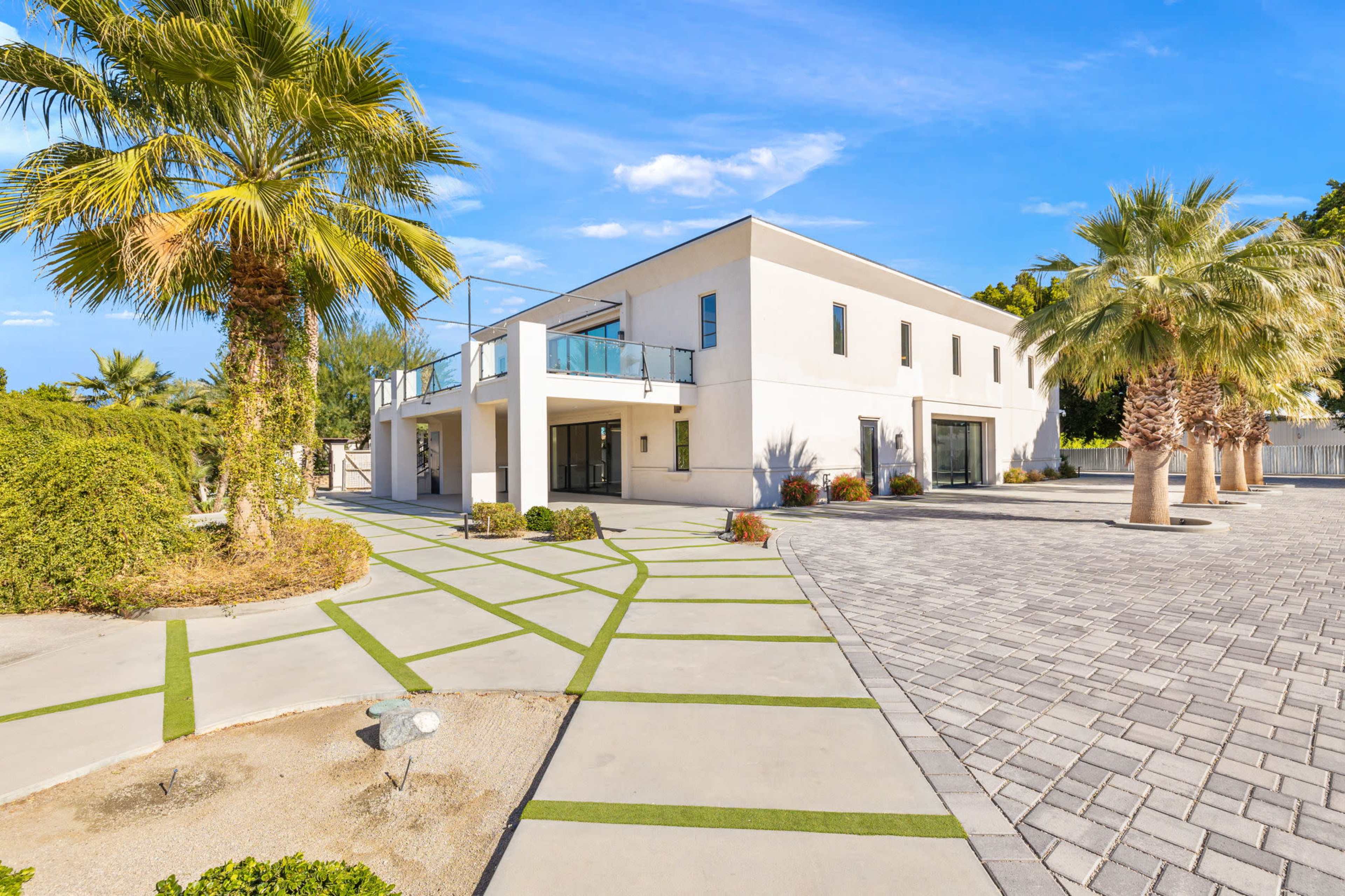 The image shows a modern two-story house surrounded by palm trees and a landscaped driveway with patterned concrete.