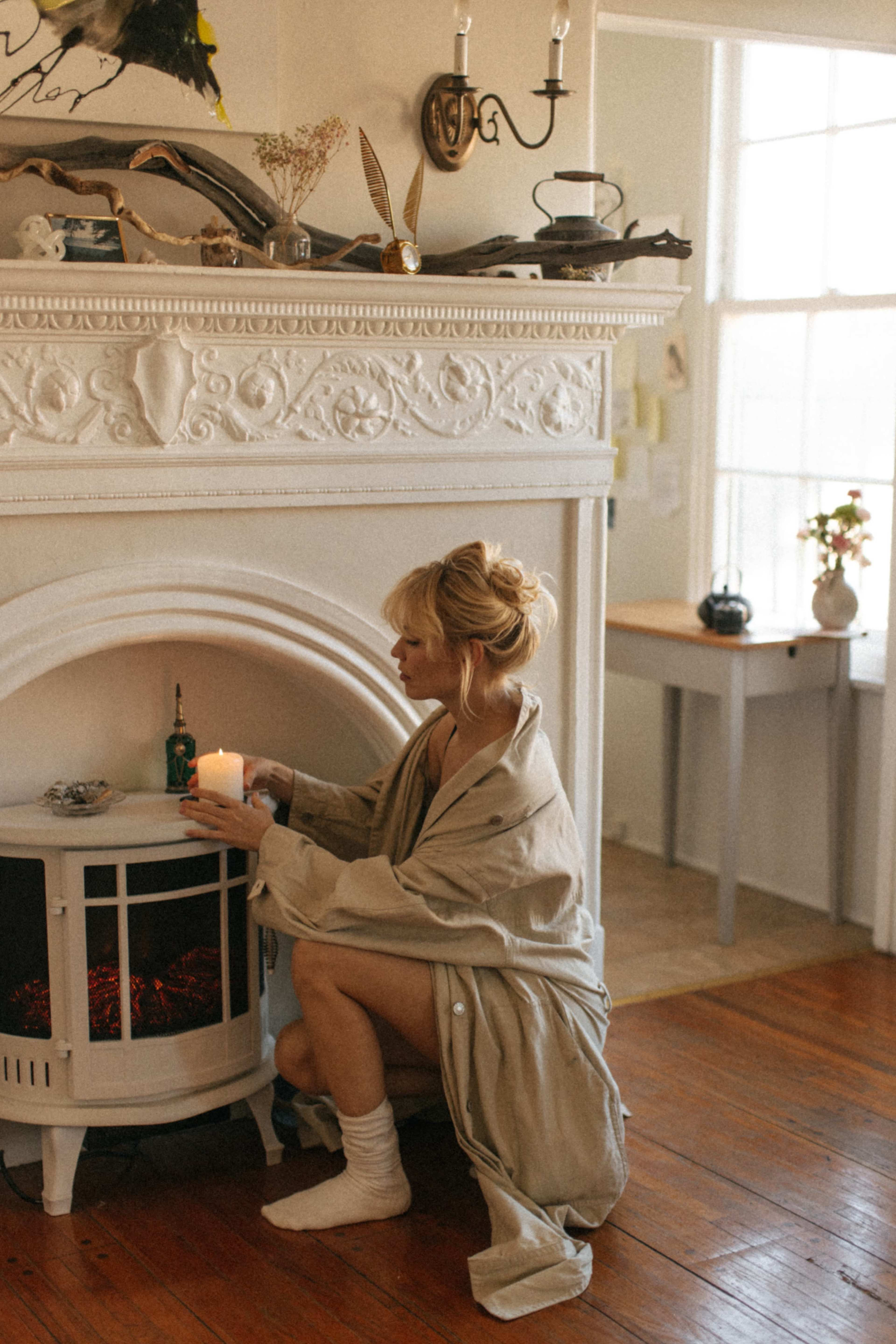 A woman in a robe kneels beside a decorative fireplace, holding a candle in a sunlit room.
